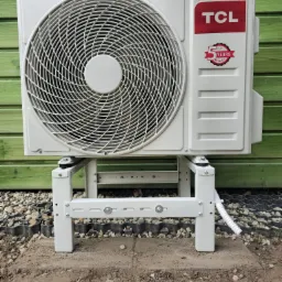 Biała jednostka zewnętrzna klimatyzacji TCL na metalowym stelażu, widoczna na tle zielonej, drewnianej ściany i podłoża z kamieni. Na urządzeniu naklejka z 5-letnią gwarancją.