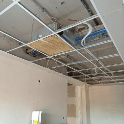KajMar Arkadiusz Rutkowski - Montaż klimatyzacji kasetonowej w biurze: widok na sufit podwieszany z widoczną jednostką wewnętrzną, instalacją elektryczną i rurami chłodniczymi. Etap prac wykończeniowych.