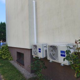 KajMar Arkadiusz Rutkowski - Dwie białe jednostki zewnętrzne klimatyzacji Haier, zamontowane na jasnobeżowej elewacji domu, z białą rurą odpływową i zielonymi krzewami róż pod spodem.