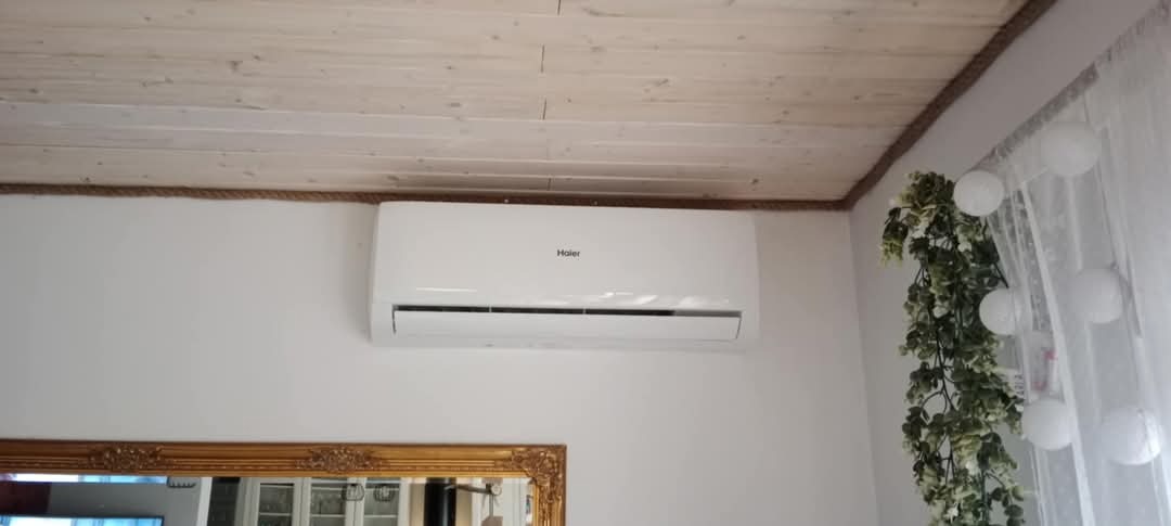 Biały klimatyzator Haier zamontowany na ścianie w pomieszczeniu z drewnianym sufitem i dekoracyjnym sznurem wzdłuż krawędzi, widoczny fragment lustra w złotej ramie i dekoracja roślinna przy oknie.