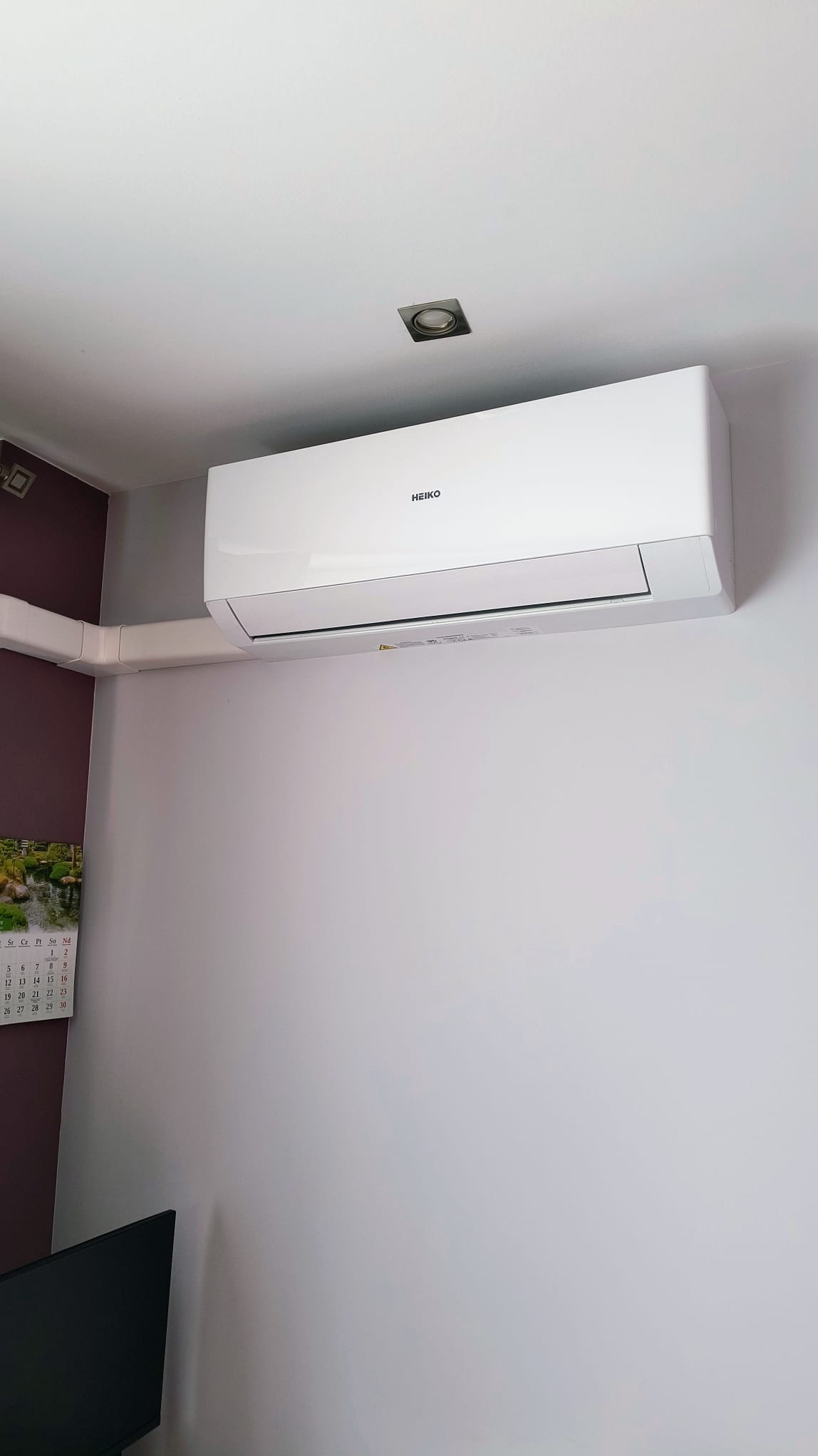 Biała jednostka klimatyzacji marki Heiko zamontowana na ścianie w pomieszczeniu, z widocznymi białymi rurami odprowadzającymi, kalendarzem na ścianie i fragmentem telewizora w dolnym rogu.