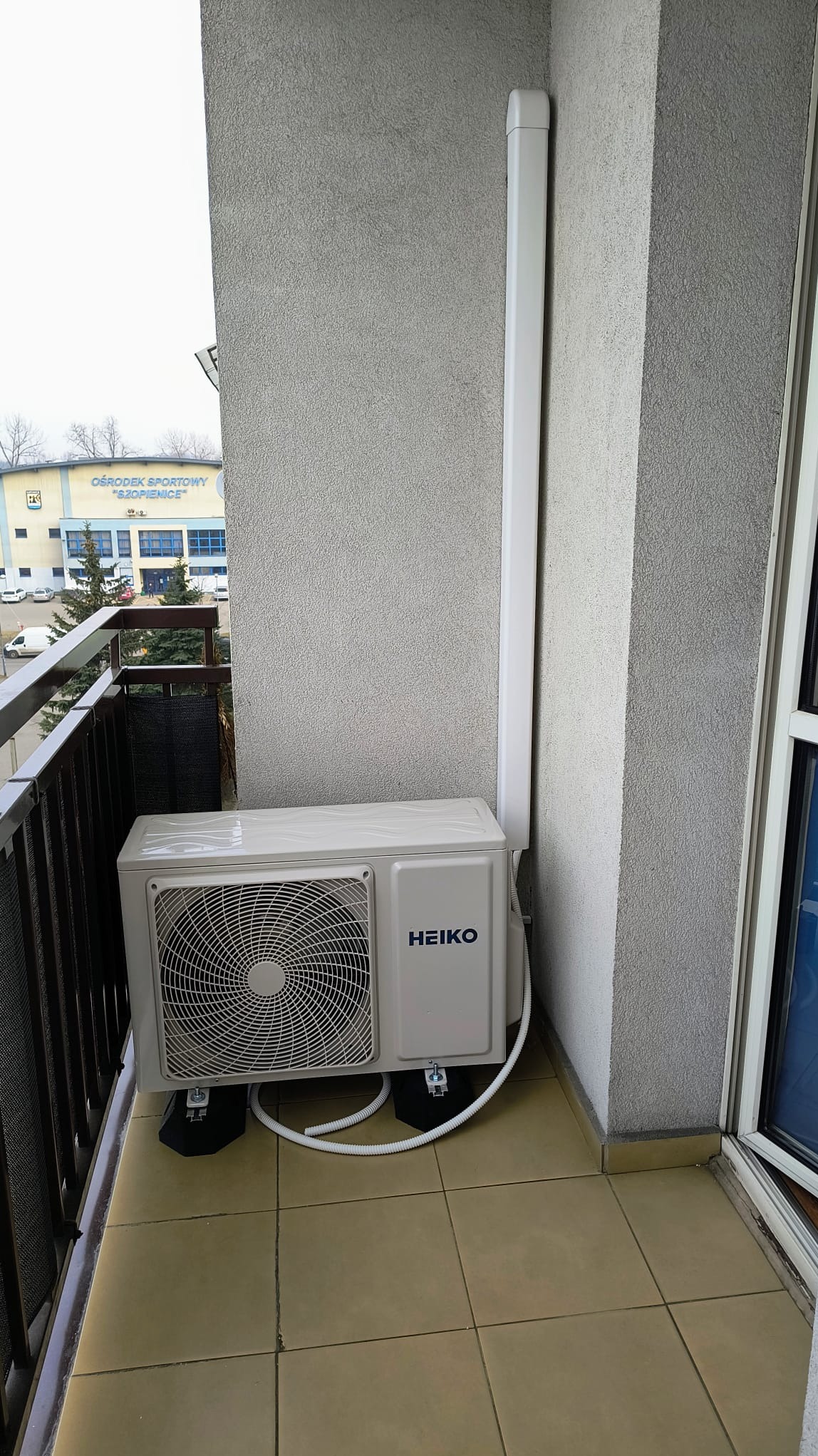 Zewnętrzna jednostka klimatyzacji Heiko zamontowana na balkonie mieszkania, widoczny budynek Ośrodka Sportowego Szopienice w tle.