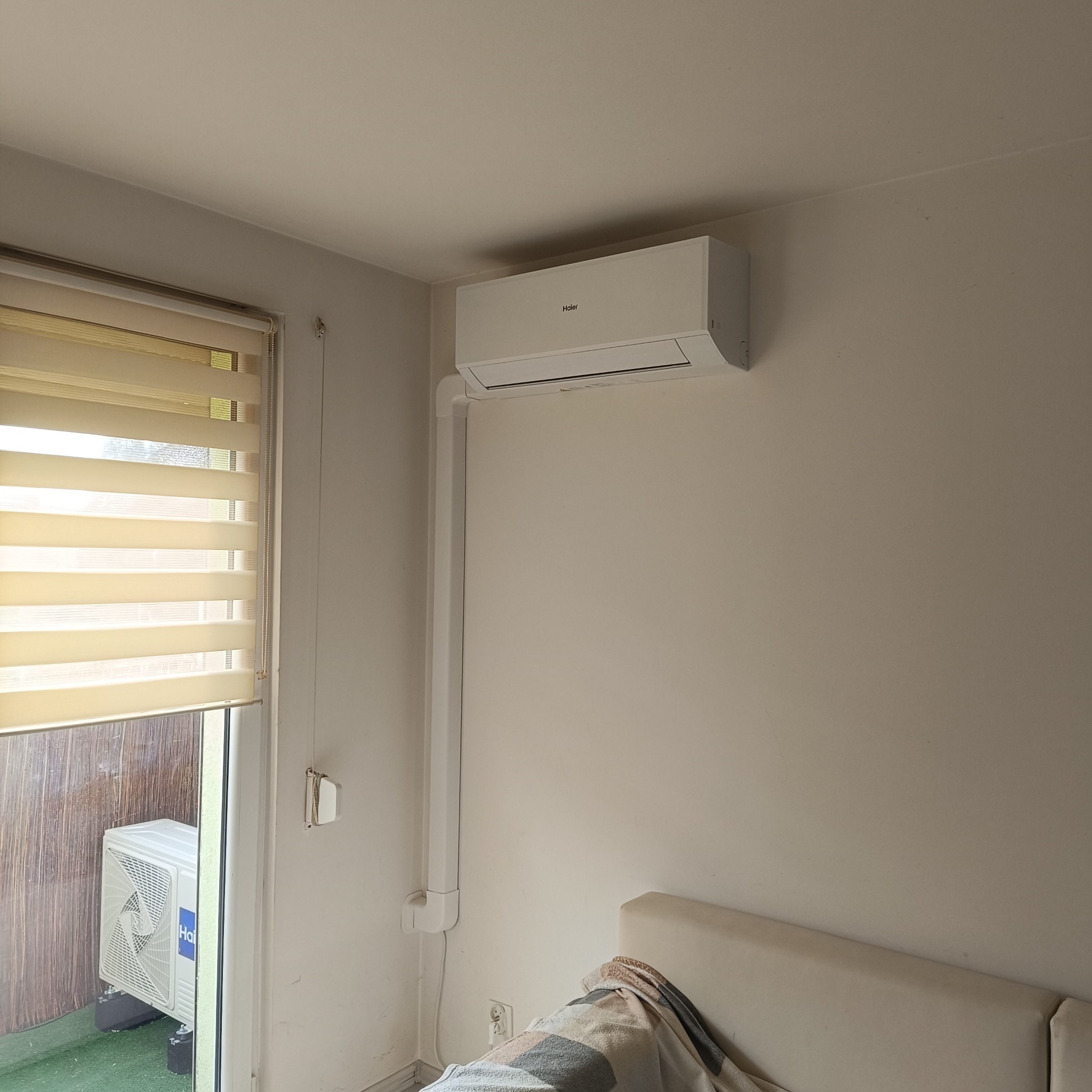 Zamontowana jednostka wewnętrzna klimatyzatora Haier na jasnej ścianie w pokoju, widoczna instalacja elektryczna w białej listwie, za oknem jednostka zewnętrzna.