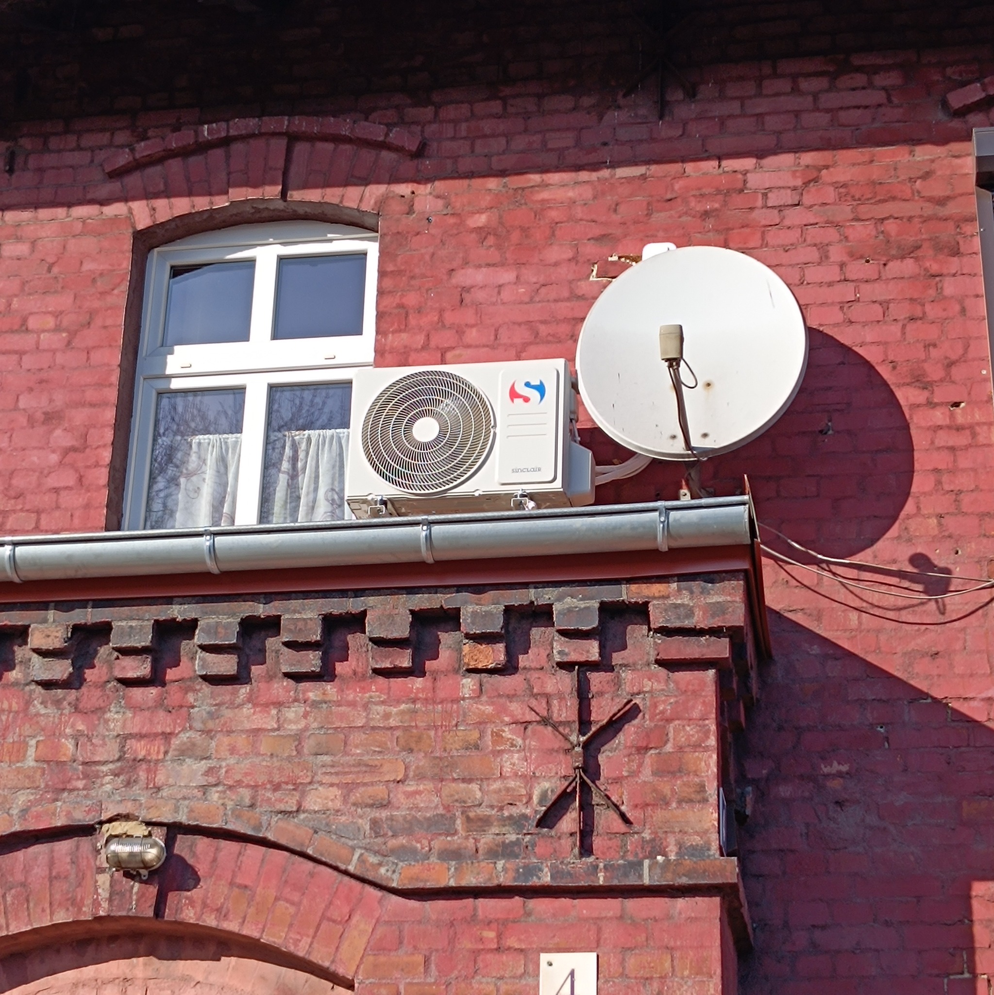 Klimatyzator Sinclair zamontowany na czerwonej ceglanej ścianie budynku obok okna i anteny satelitarnej.