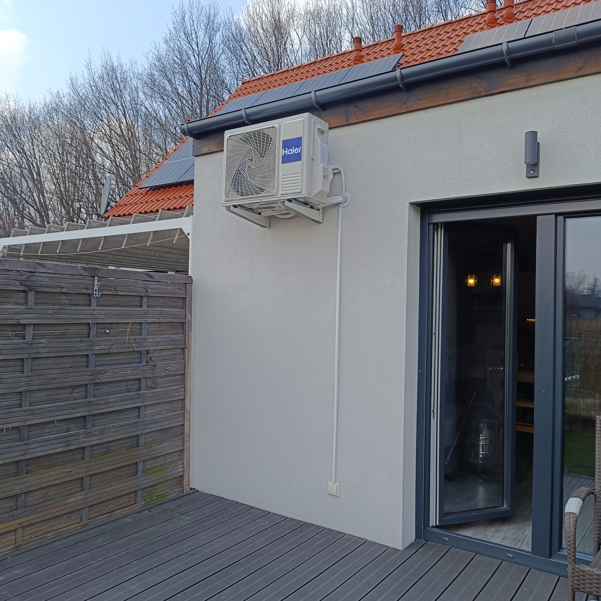 Zewnętrzna jednostka klimatyzacji Haier zamontowana na ścianie domu z panelami słonecznymi na dachu i otwartymi drzwiami balkonowymi.
