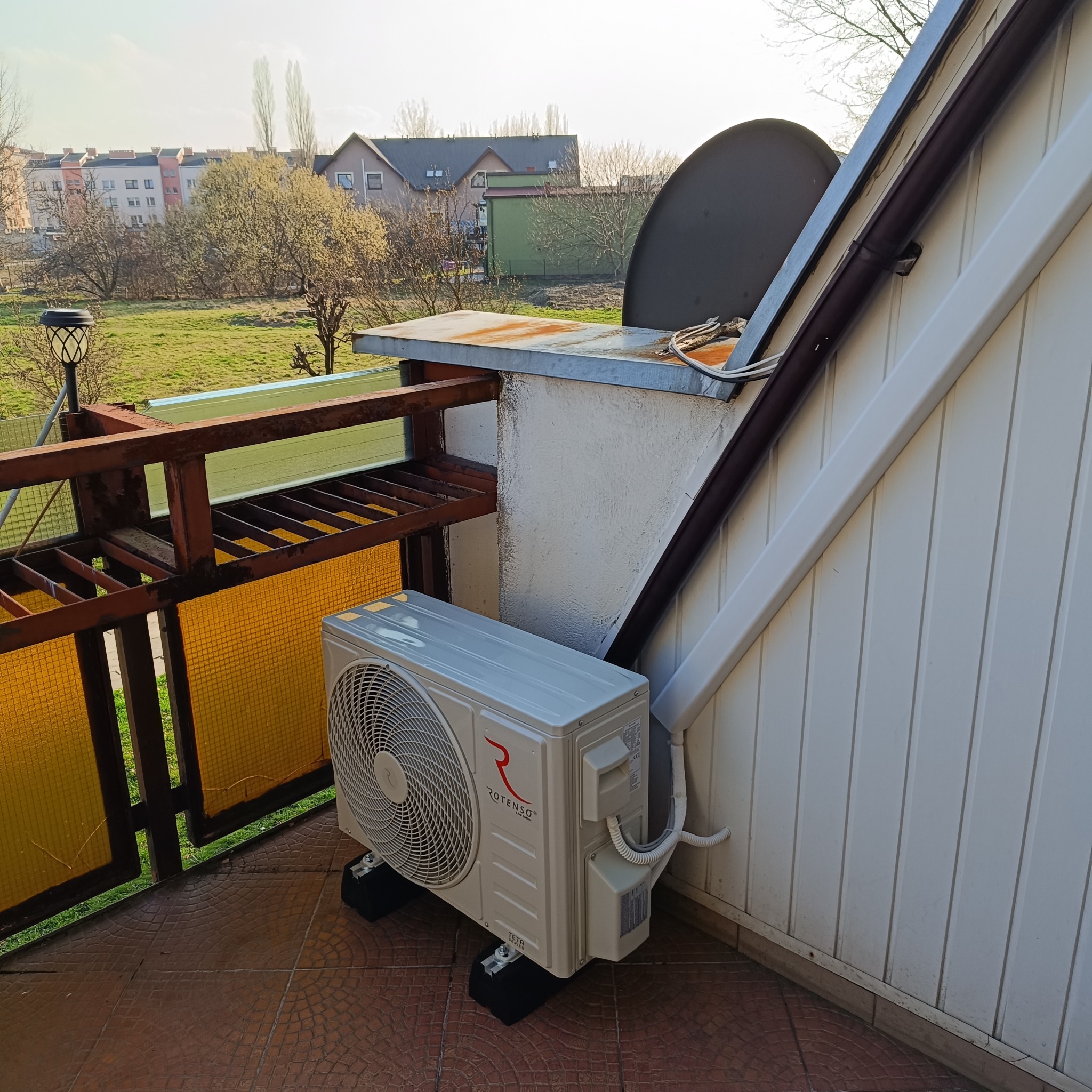 Biała jednostka zewnętrzna klimatyzacji Rotenso zamontowana na balkonie, widok na osiedle mieszkaniowe w tle.