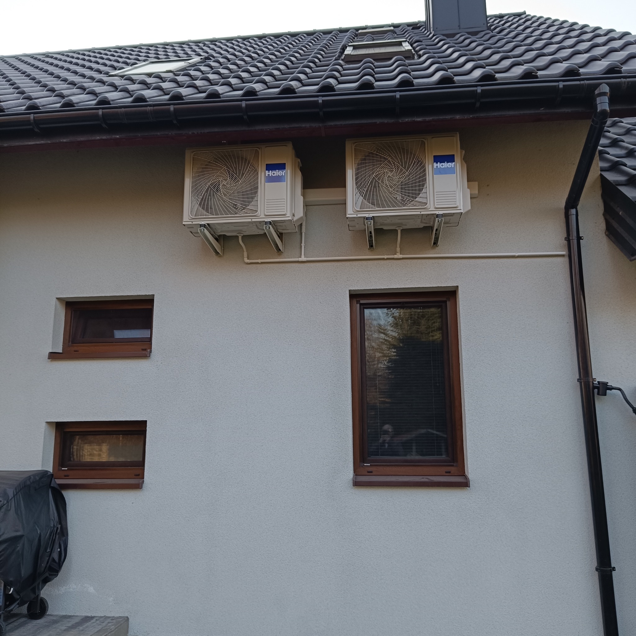 Dwa klimatyzatory marki Haier zamontowane na elewacji domu, widoczne okna, rynna i fragment dachu z dachówką ceramiczną.