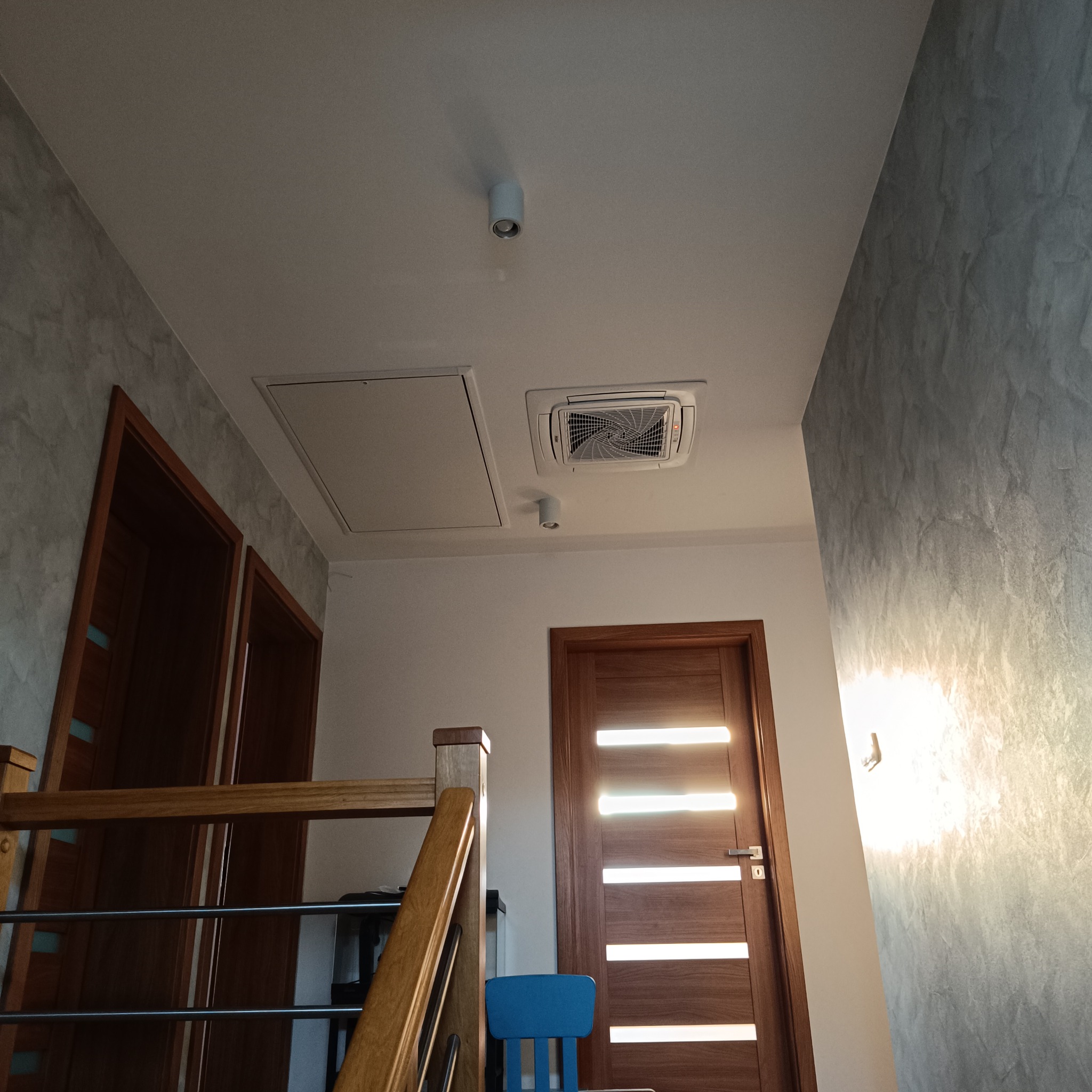 Wnętrze domu z zamontowaną kasetonową jednostką klimatyzacji sufitowej, widok spod schodów na korytarz z dwiema parami drzwi i oświetleniem punktowym.