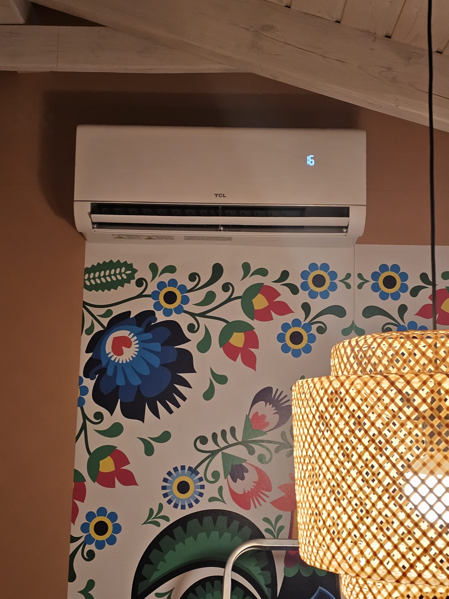 Biały klimatyzator TCL zamontowany na ścianie z ludowym wzorem kwiatowym, widoczny fragment lampy z rattanu, temperatura ustawiona na 16 stopni Celsjusza.
