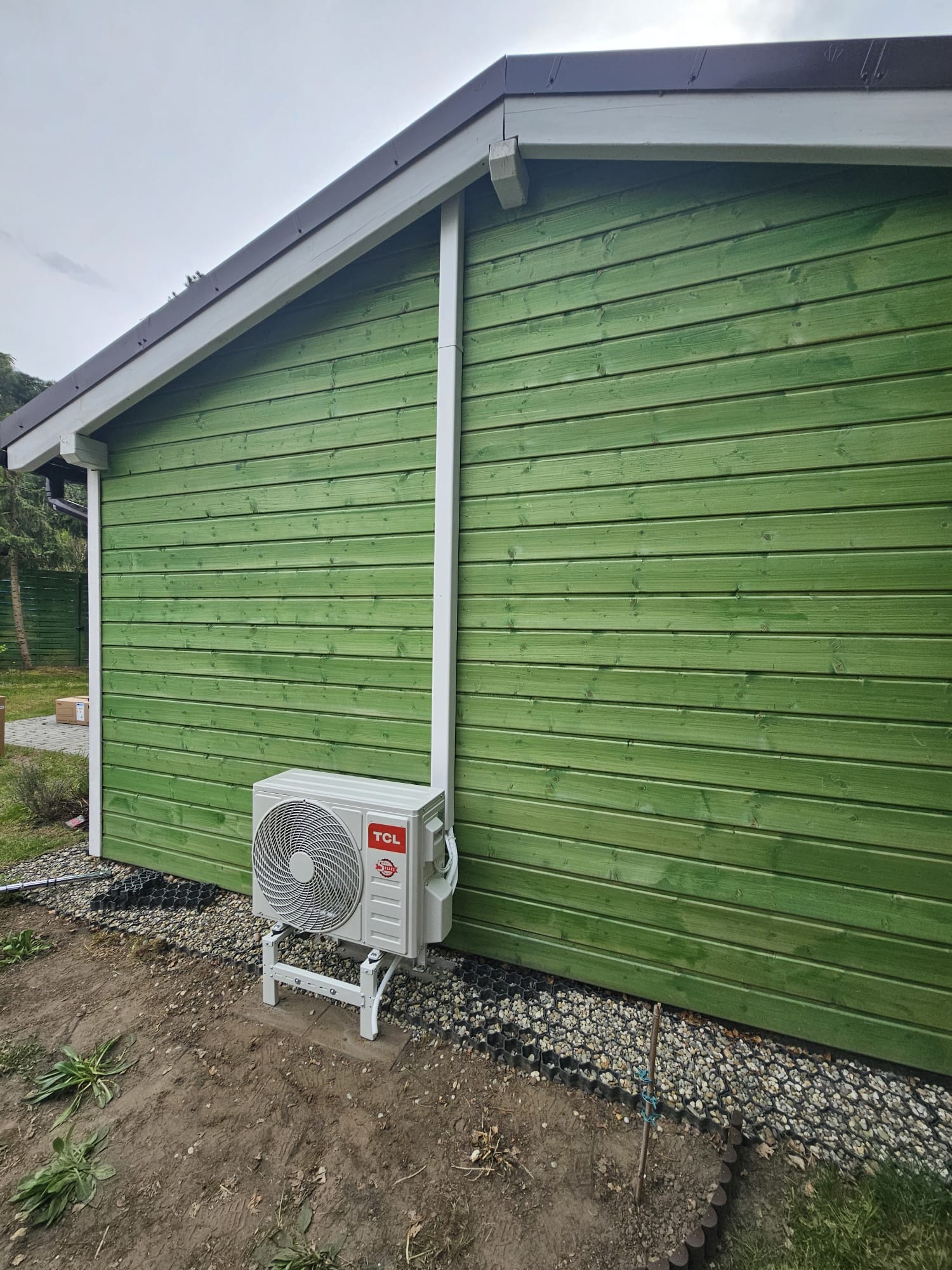 Biały klimatyzator TCL zamontowany na zewnątrz zielonego, drewnianego domku z brązowym dachem, z widocznymi elementami instalacji i otoczeniem z ziemią i kamieniami.