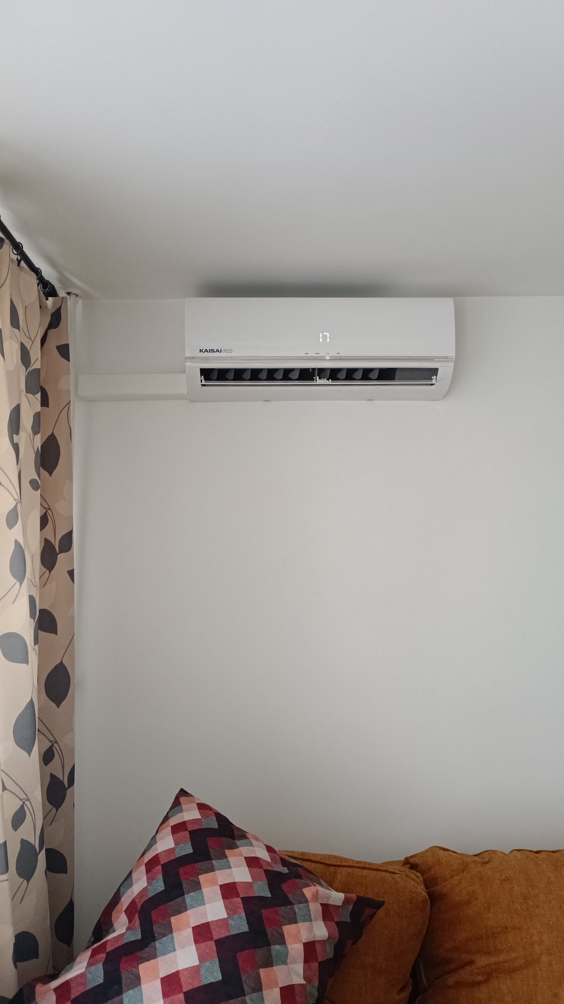 Biały klimatyzator Kaisai Eco z wyświetlaczem temperatury (17°C) zamontowany na ścianie w Katowicach. Widoczny fragment zasłony i poduszki na kanapie.