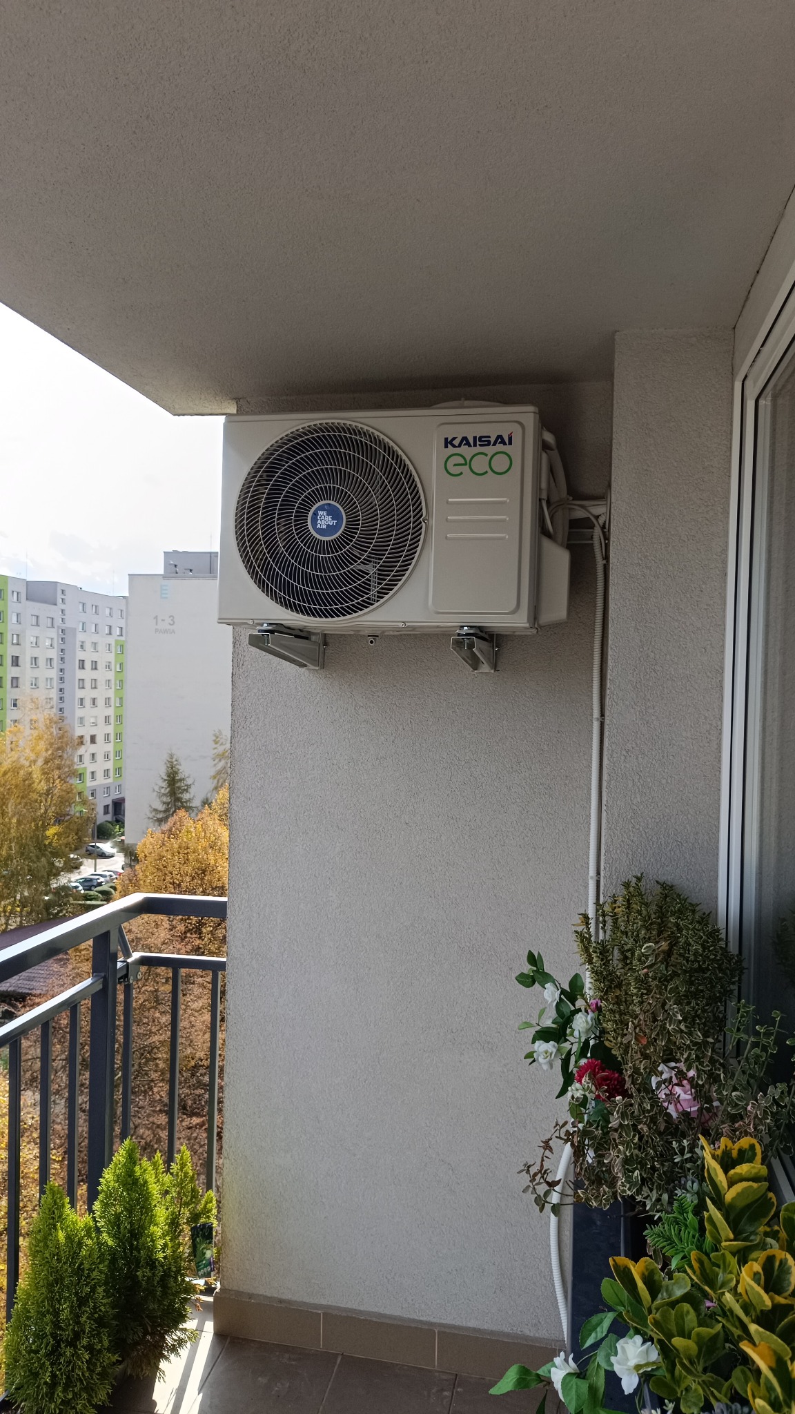 Zamontowana jednostka zewnętrzna klimatyzacji Kaisai Eco na balkonie mieszkania w bloku. Widoczne przewody i rośliny w donicach.