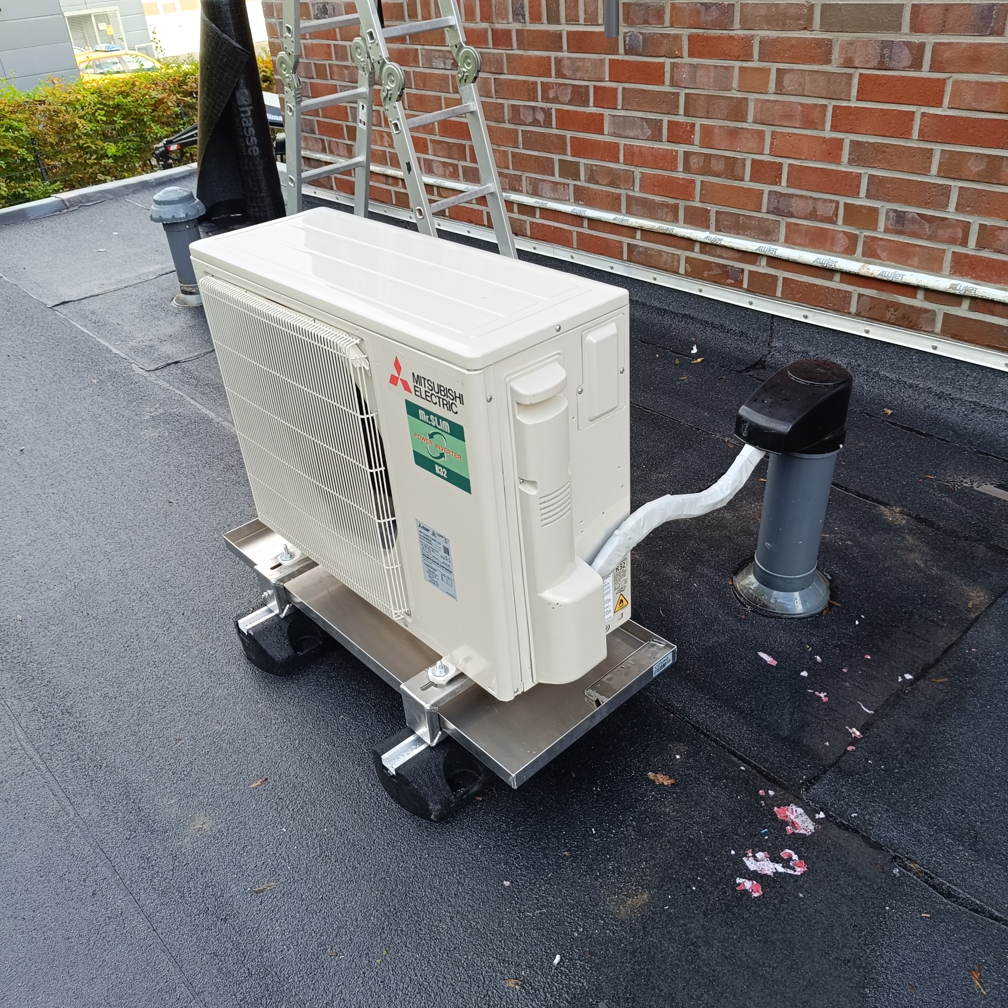 Zamontowana jednostka zewnętrzna klimatyzacji Mitsubishi Electric na dachu, widoczna drabina i komin wentylacyjny. Instalacja na metalowej podstawie z amortyzatorami.