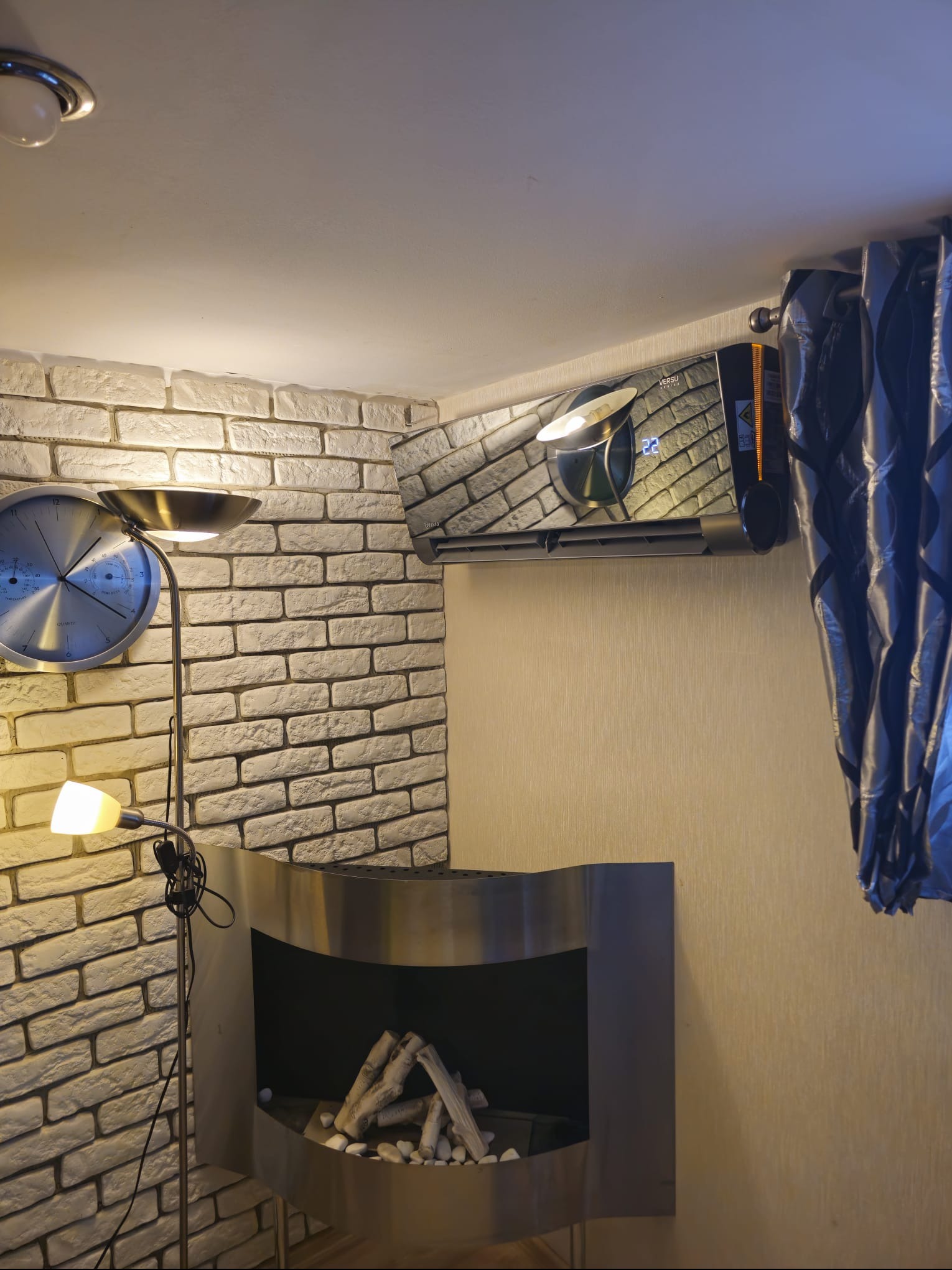 Klimatyzator ścienny w nowoczesnym wnętrzu z ceglaną ścianą, lampą podłogową i dekoracyjnym kominkiem. Widoczny wyświetlacz temperatury i zasłony w tle.