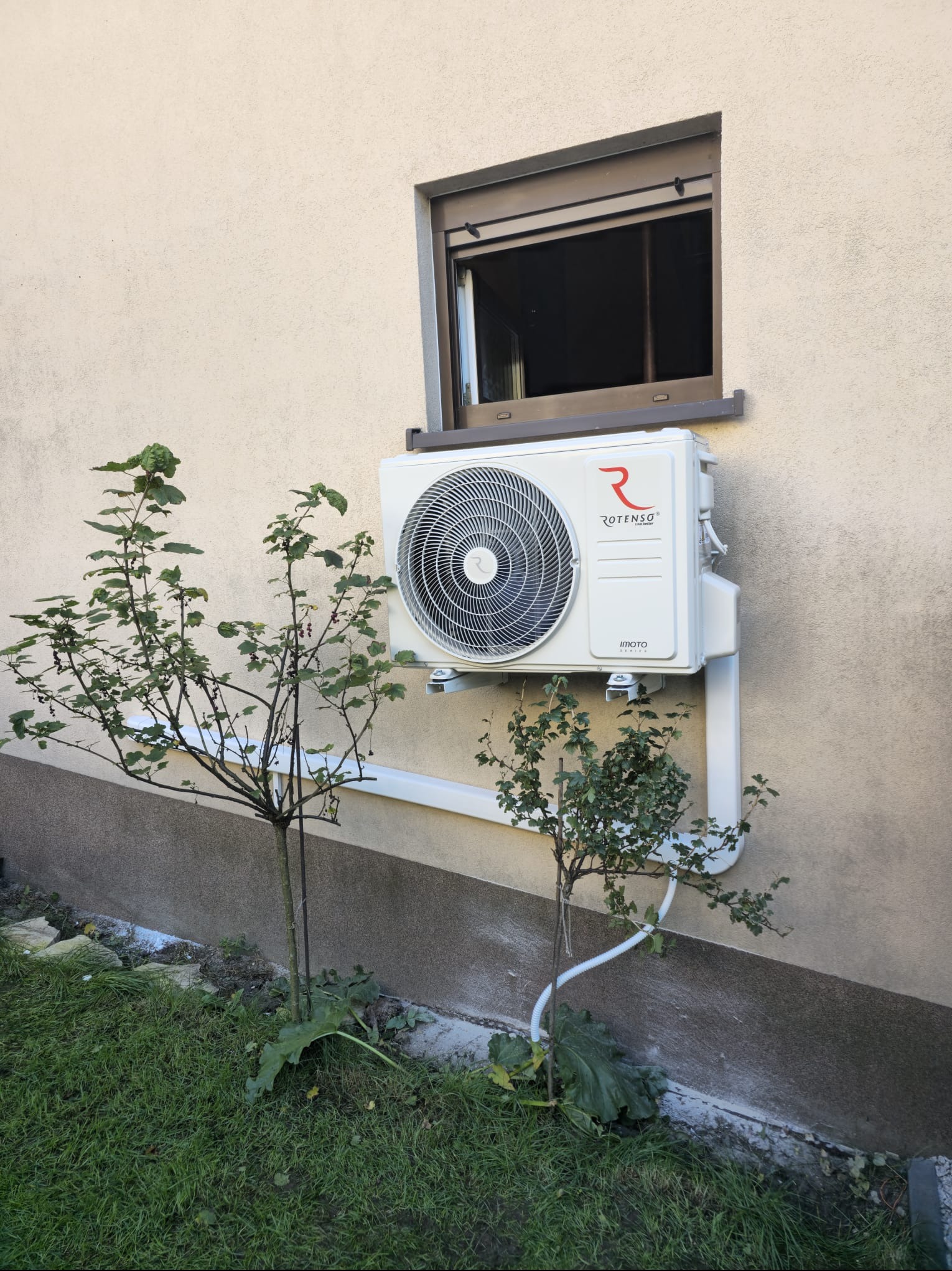 Biały klimatyzator Rotenso zamontowany na elewacji domu pod oknem, z widoczną instalacją odprowadzającą skropliny, otoczony zielenią. Ujęcie z dołu.