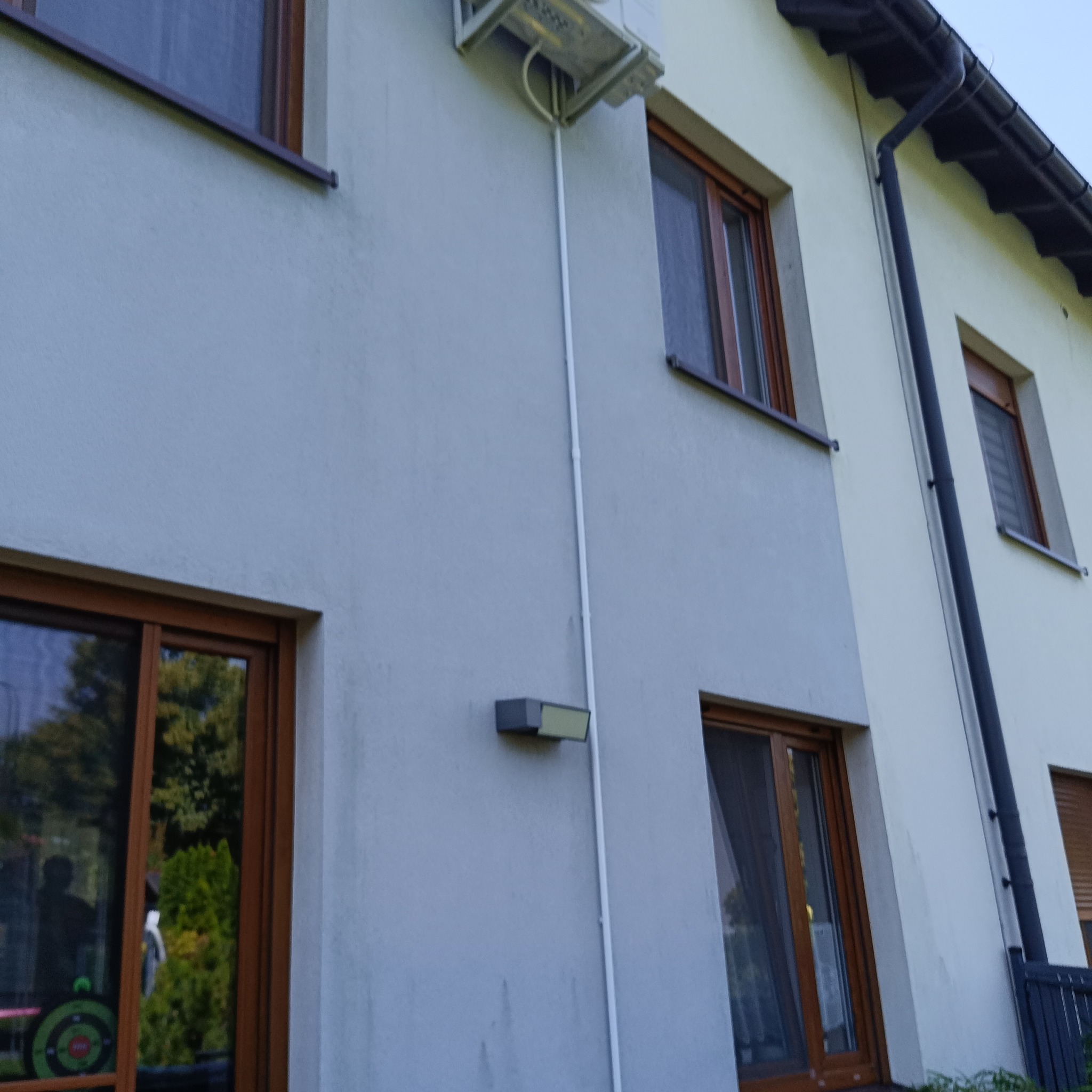 Biała elewacja domu z zamontowanym klimatyzatorem. Widoczne okna z brązowymi ramami i biała rura instalacyjna. Ujęcie z dołu, lekko zniekształcające perspektywę.