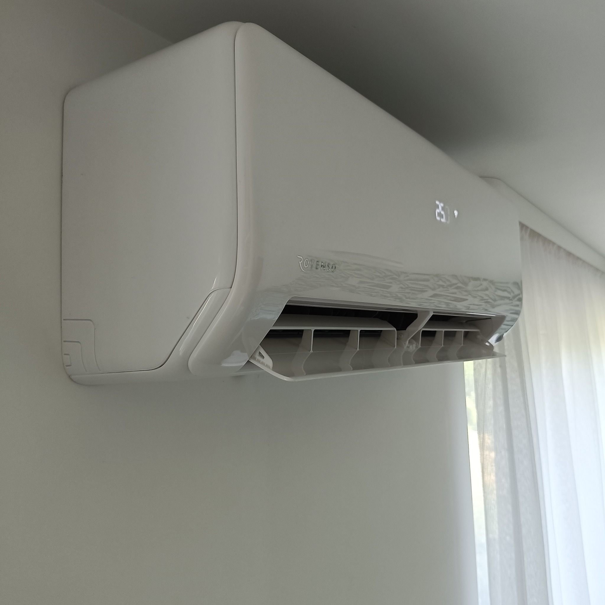 Biała klimatyzacja ścienna Rotenso z wyświetlaczem 25 stopni Celsjusza, zamontowana w jasnym pomieszczeniu z zasłoną w tle.
