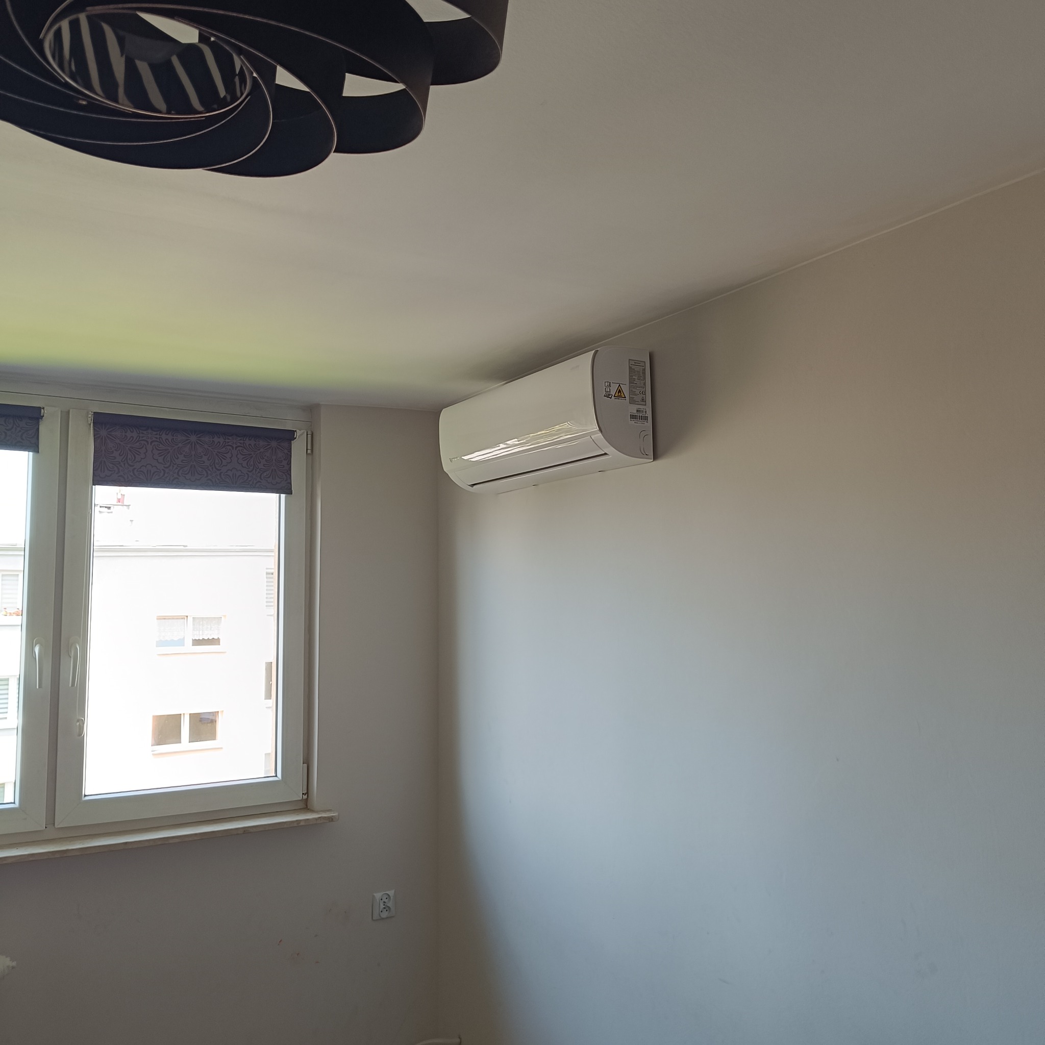 Biały klimatyzator ścienny w jasnym pokoju z oknem i roletami. Widoczny fragment lampy sufitowej. Minimalistyczny design, neutralne kolory ścian.