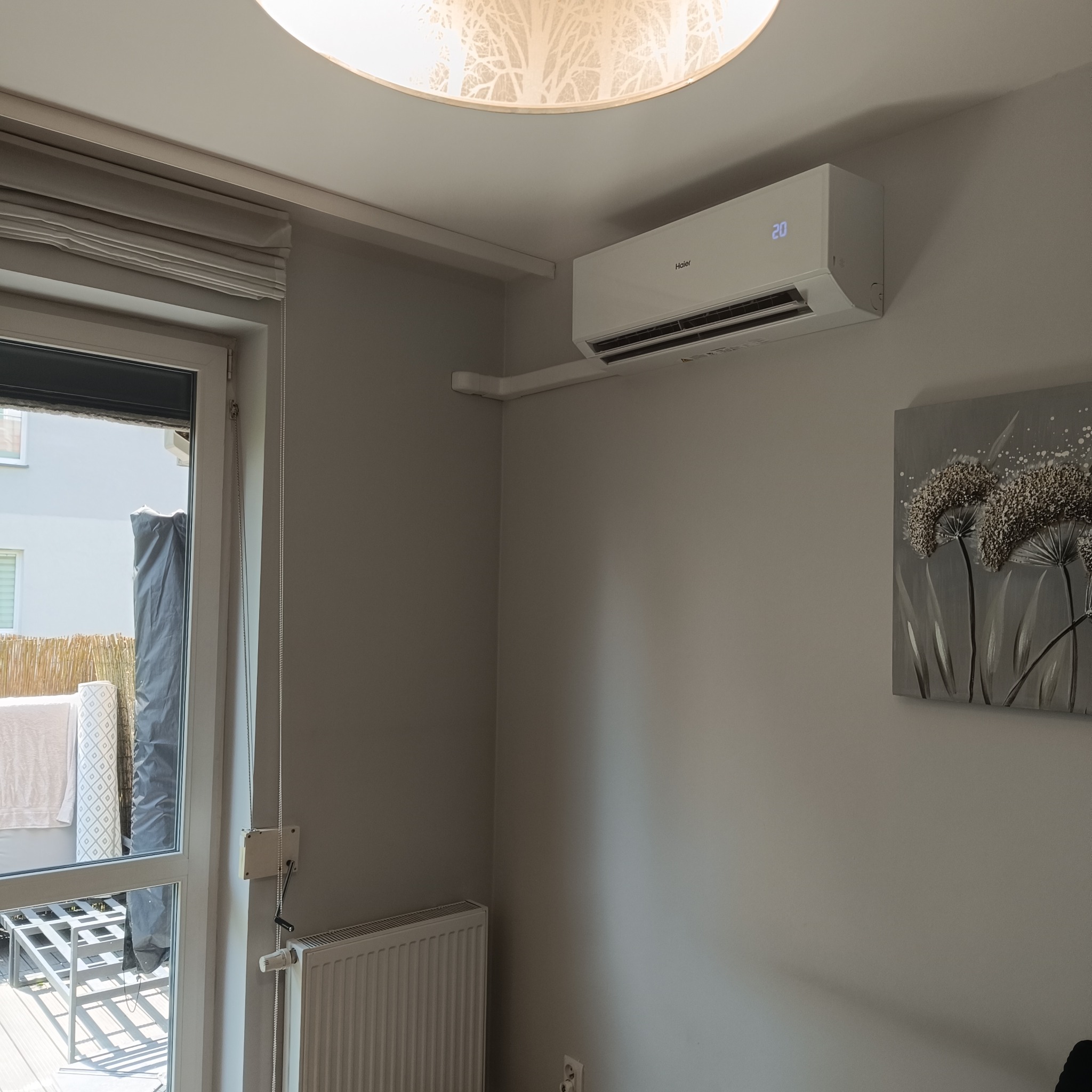 Biały klimatyzator Haier z wyświetlaczem temperatury (20°C) zamontowany na szarej ścianie w pokoju z obrazem kwiatów i widokiem na balkon.