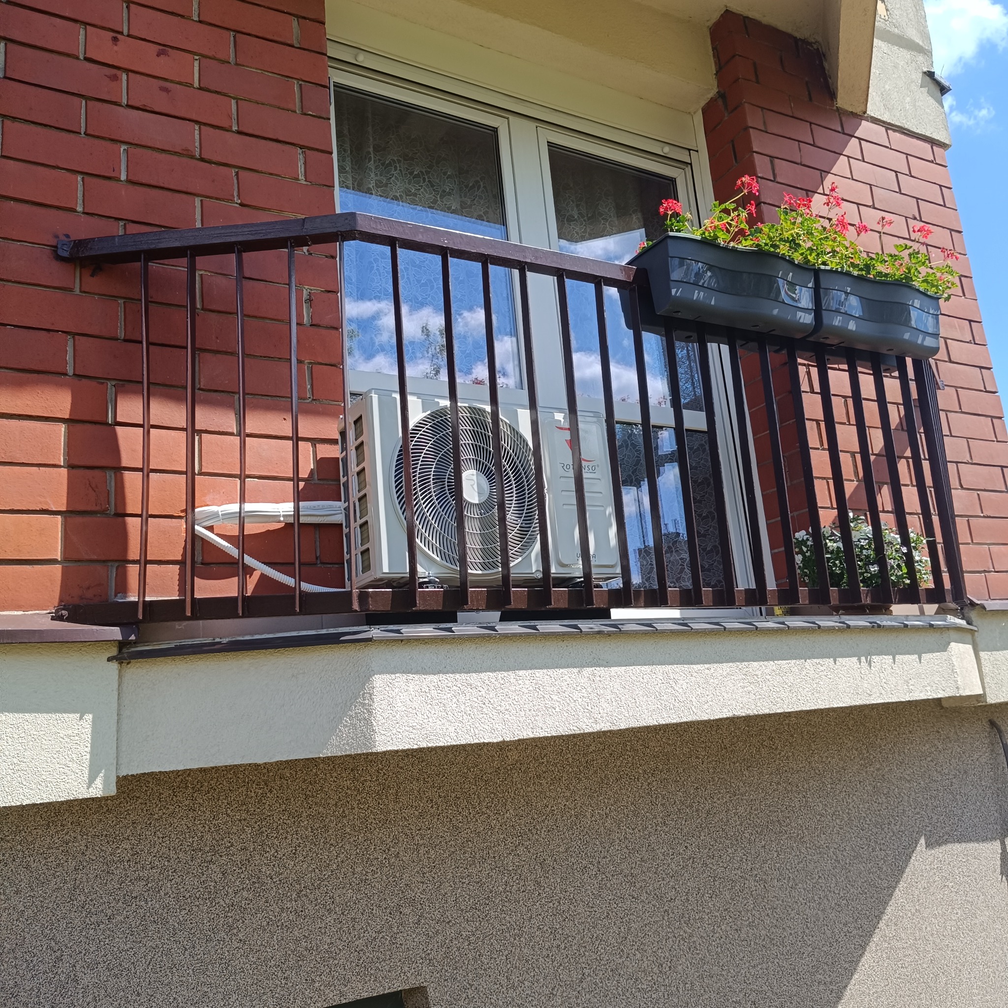 Biała jednostka klimatyzacji na balkonie z ceglaną ścianą, widoczna przez metalową balustradę, z kwiatami w donicach. Instalacja na zewnątrz budynku.