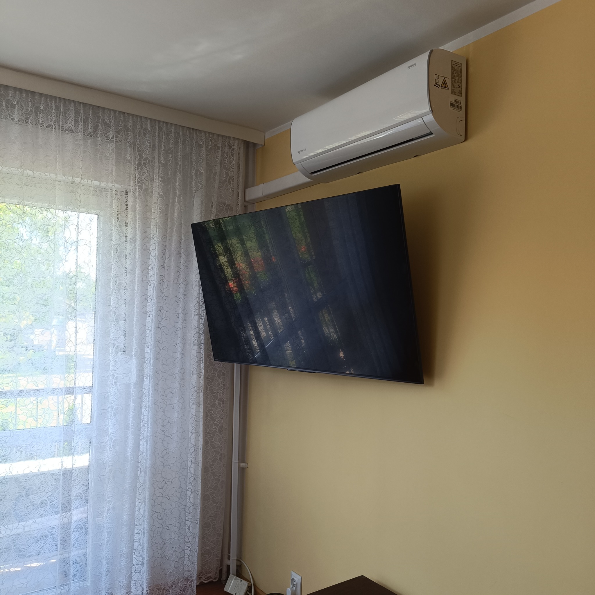 Biały klimatyzator ścienny zamontowany nad telewizorem na żółtej ścianie w pokoju z oknem i firanką. Widoczne kable w listwie.