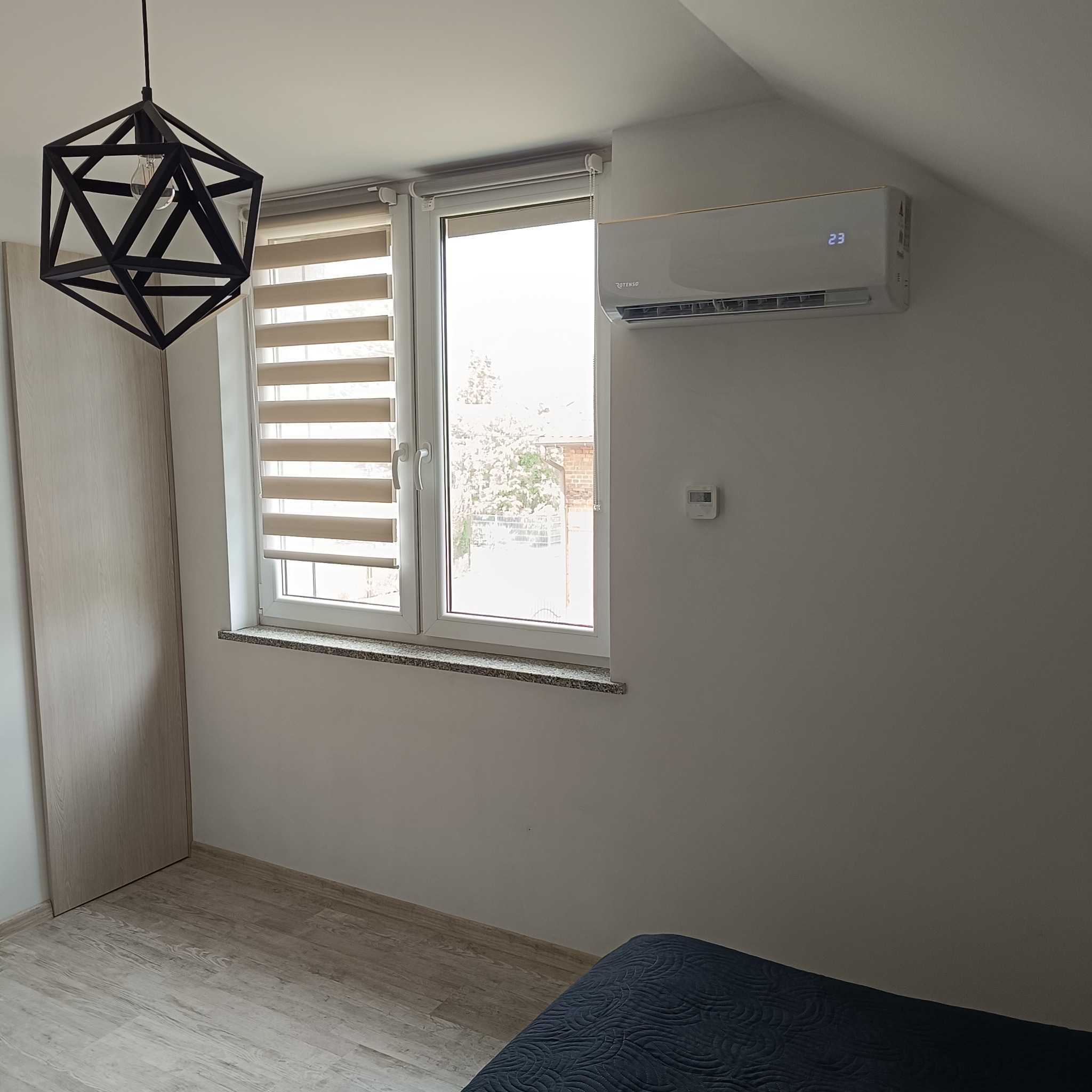 Biała klimatyzacja Rotenso zamontowana nad oknem z żaluzjami w jasnym pokoju. Widoczny geometryczny żyrandol i fragment łóżka. Temperatura na wyświetlaczu klimatyzacji to 23 stopnie.