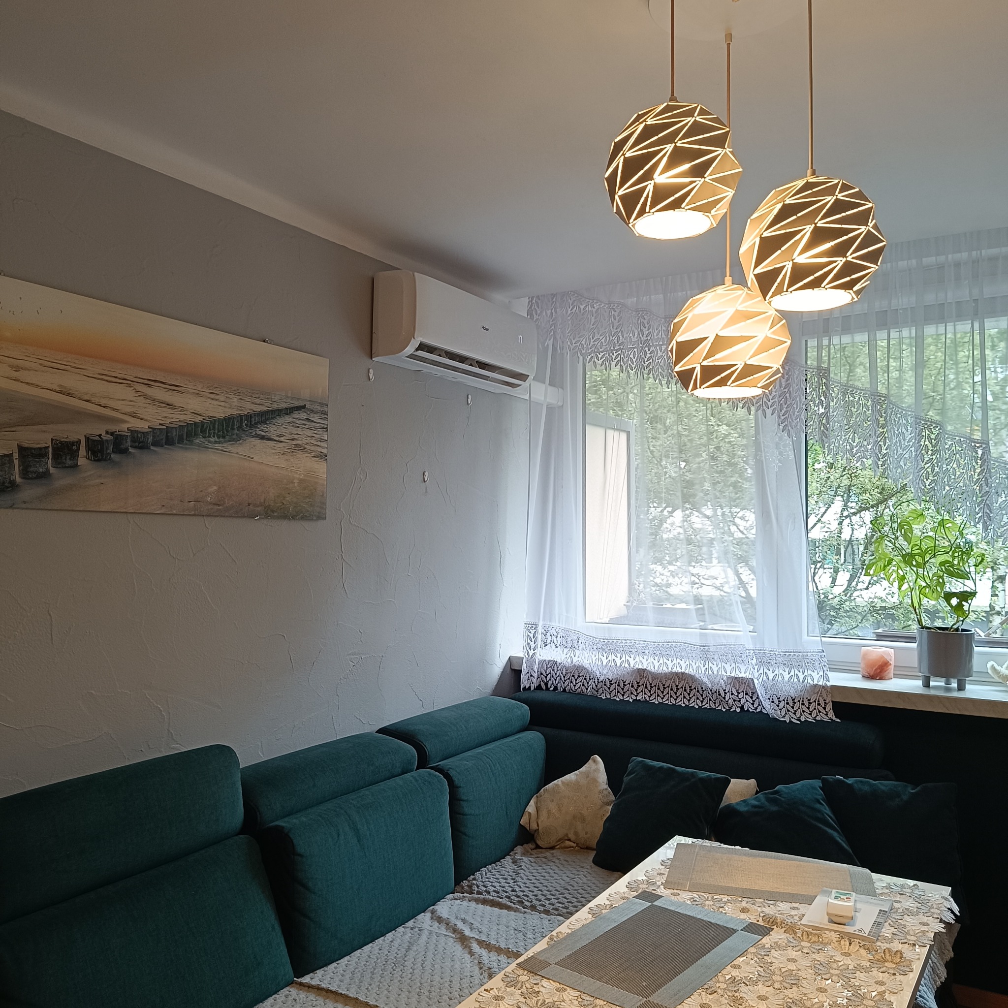 Nowoczesny klimatyzator Haier na ścianie w salonie z zieloną sofą, widokiem z okna i designerskimi lampami. Estetyczne wnętrze.