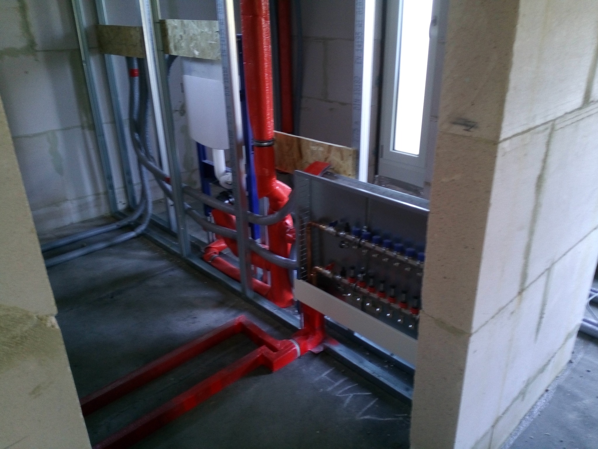 Surowy stan instalacji podłogowej z czerwonymi rurami w metalowej ramie, widoczna rozdzielnica z licznymi zaworami, ściany z betonu komórkowego w trakcie budowy.