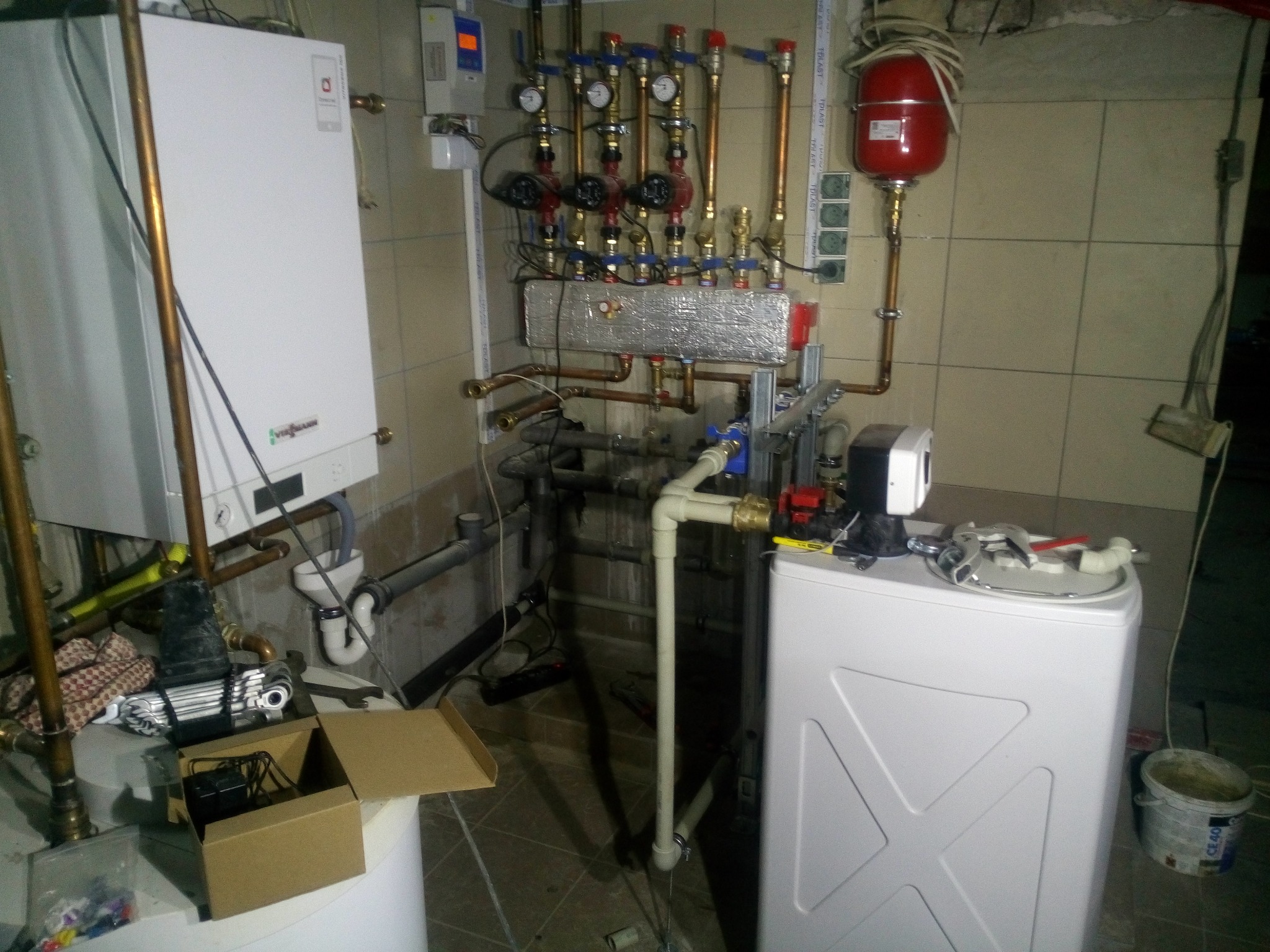 Złożona instalacja kotła grzewczego z licznymi rurami miedzianymi, zaworami i manometrami, widoczny biały kocioł oraz czerwony zbiornik wyrównawczy, całość w pomieszczeniu z kafelkami.