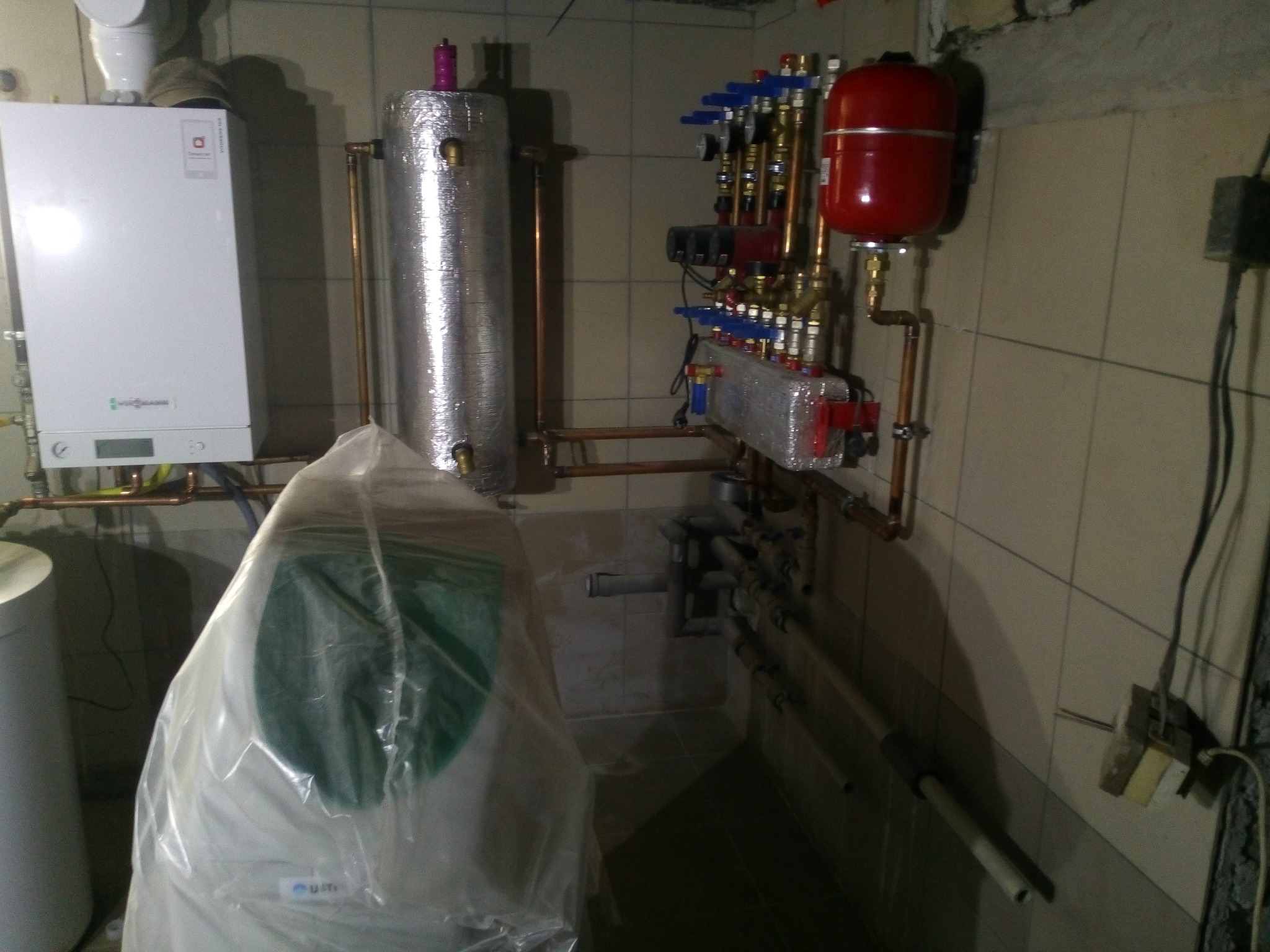 Kotłownia z piecem Viessmann, zasobnikiem ciepłej wody pod folią ochronną, zbiornikiem wyrównawczym i rozdzielaczem z pompami obiegowymi na tle kafelkowanej ściany.