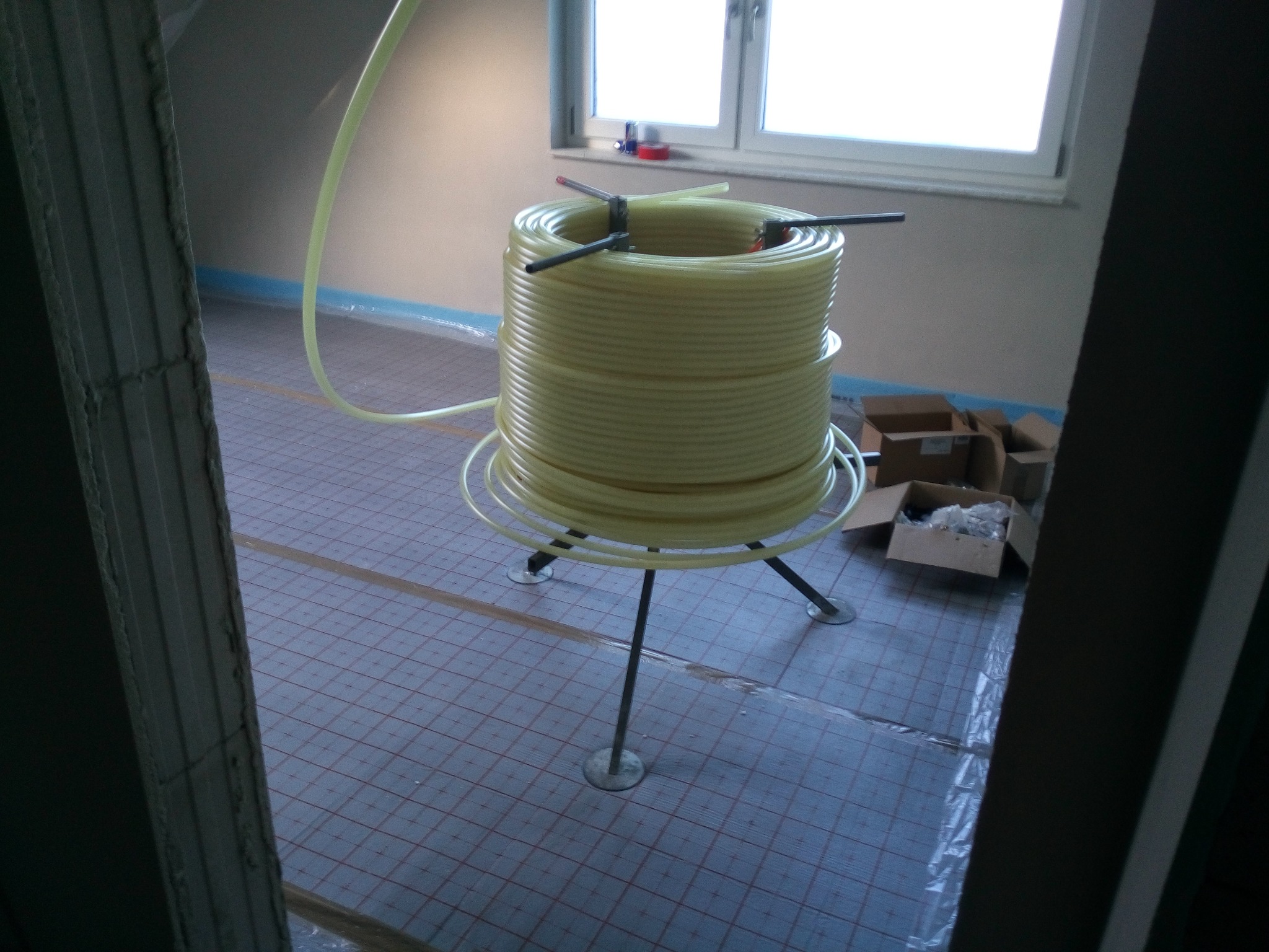 Żółta rura zwinięta w krąg na metalowym stojaku podczas montażu ogrzewania podłogowego, podłoga zabezpieczona folią, widoczne kartony w tle.