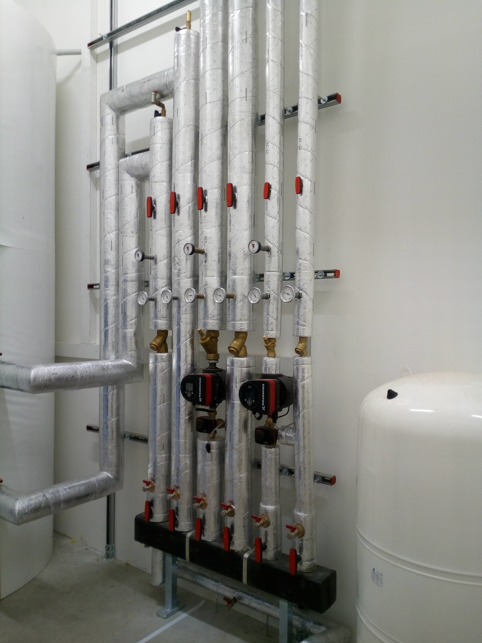 Zestaw pionowych rur w izolacji aluminiowej z czerwonymi zaworami i manometrami, podłączonych do dwóch pomp obiegowych Grundfos oraz zbiornika wyrównawczego w kotłowni.