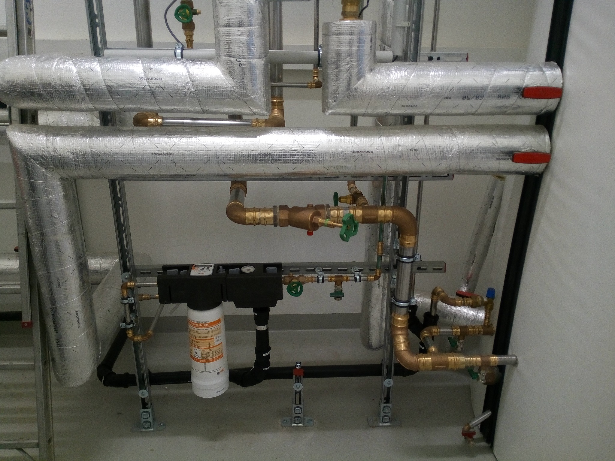Kompleksowa instalacja grzewcza z rurami izolowanymi folią aluminiową, widoczny filtr wody Solutech HRC oraz liczne zawory i złączki mosiężne.