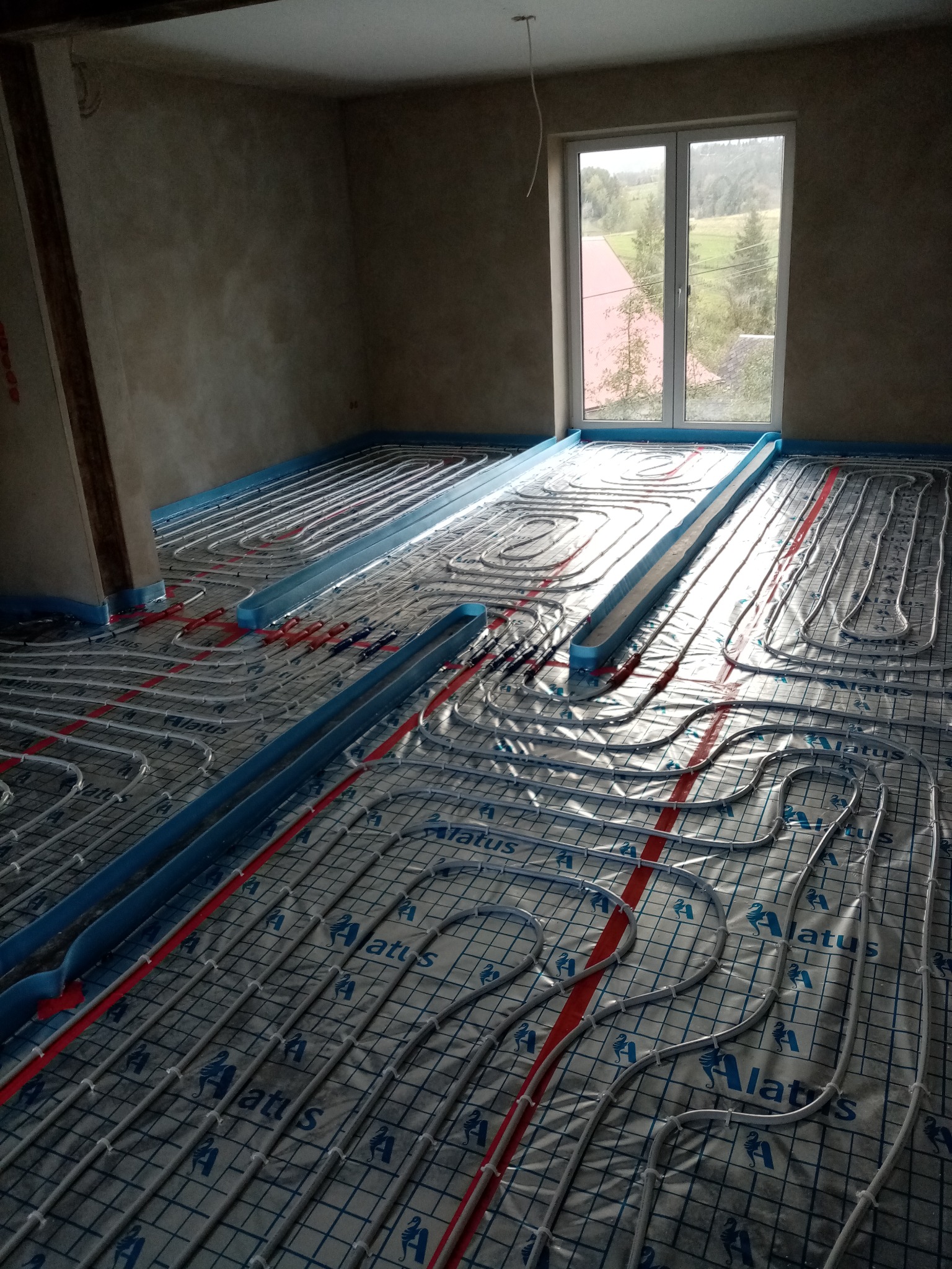 Rozłożone rury ogrzewania podłogowego na folii z nadrukiem, widoczne nieotynkowane ściany i okno z widokiem na zewnątrz.