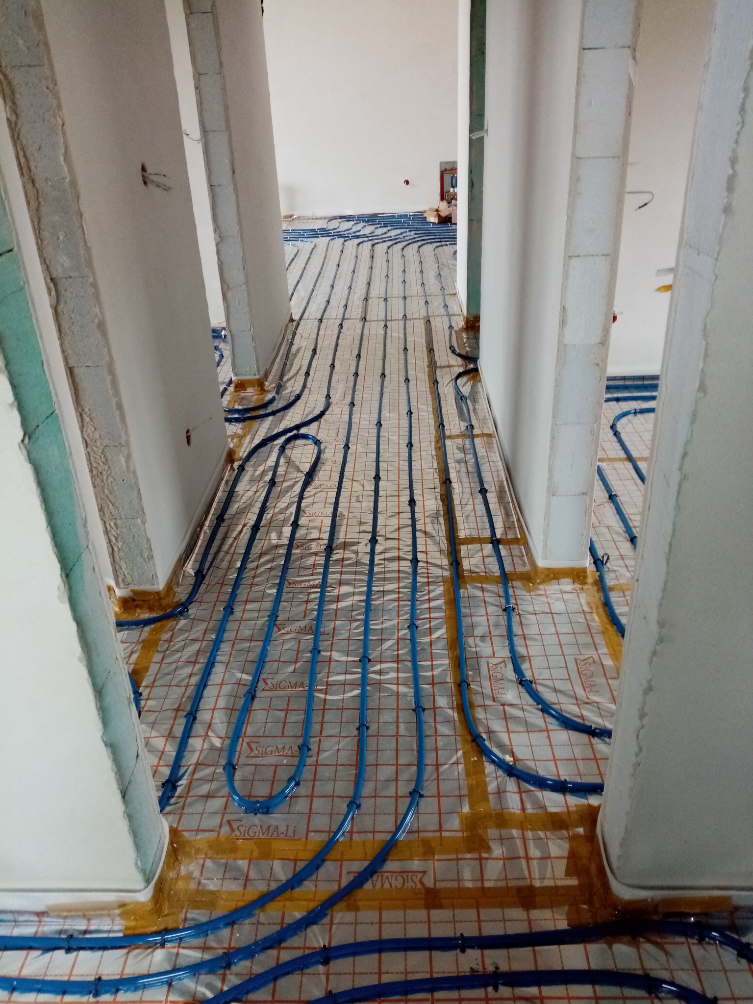 Niebieskie rury ogrzewania podłogowego ułożone na folii z siatką, zamocowane do podłoża taśmą, widoczne fragmenty ścian w surowym stanie.