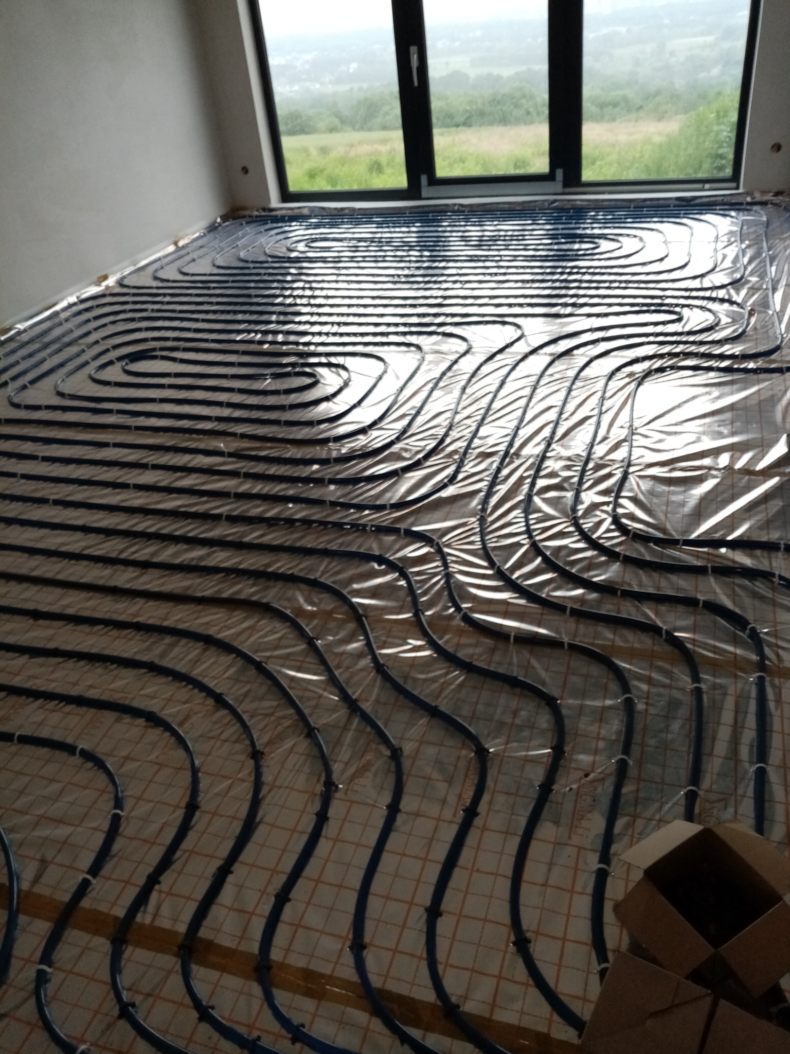 Niebieskie rurki ogrzewania podłogowego ułożone spiralnie na folii izolacyjnej z siatką w Mszanie Dolnej, widok z góry, przez okno widać zielony krajobraz.