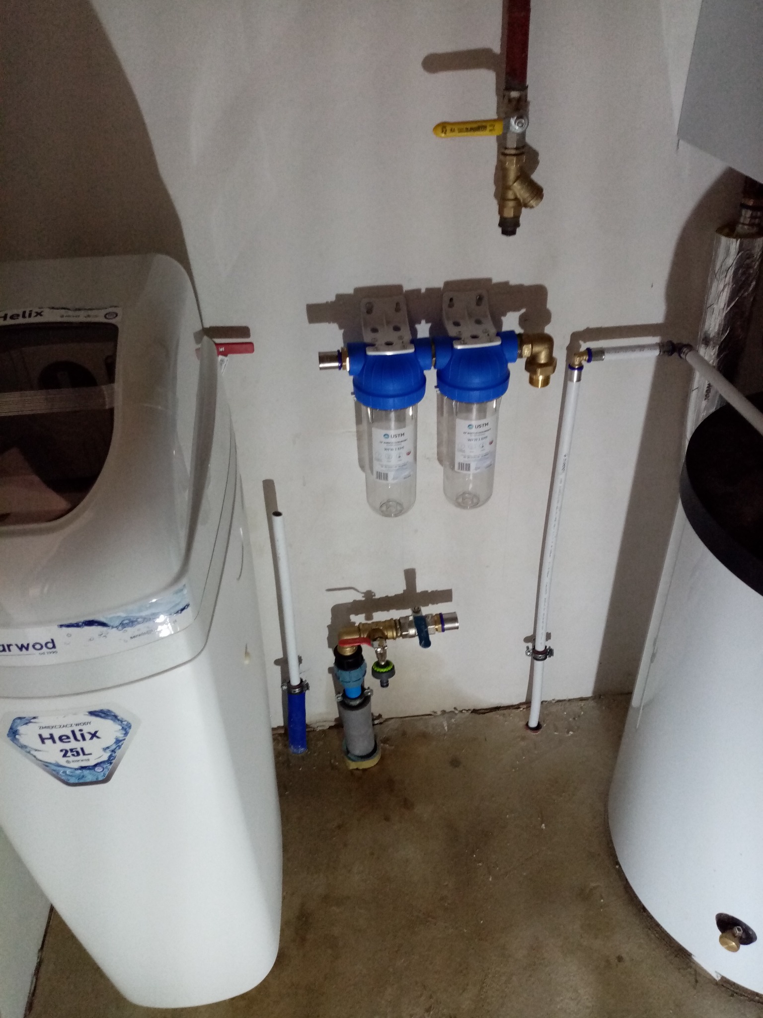 Biały zmiękczacz wody Helix 25L, dwa filtry wody z niebieskimi obudowami, zawory i rury instalacji wodnej, oraz biały bojler w pomieszczeniu o betonowej podłodze.