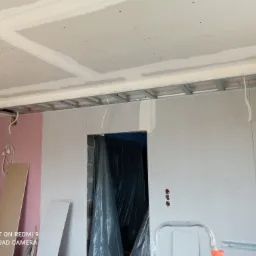 Wnętrze w trakcie montażu sufitu podwieszanego z płyt kartonowo-gipsowych, widoczne metalowe profile, częściowo położone płyty i wystające kable elektryczne. Ściany przygotowane pod dalsze prace...