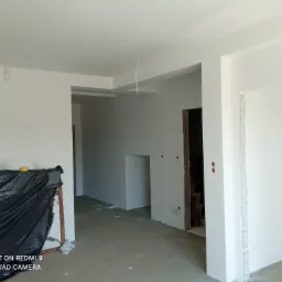 Wnętrze w trakcie prac wykończeniowych: świeżo pomalowane na biało ściany i sufit, zabezpieczona folią podłoga, widoczne przewody wystające z sufitu, ślady po gniazdkach elektrycznych w ścianach.