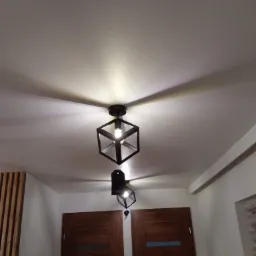 Dwie czarne lampy sufitowe w kształcie sześcianu z żarówkami, oświetlające biały sufit w pomieszczeniu z drewnianymi drzwiami i panelami ściennymi.