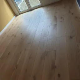 Ukończona podłoga z paneli drewnopodobnych, ułożonych równolegle do okna, z widoczną strukturą drewna i subtelnymi sękami, w jasnym pomieszczeniu z żółtą ścianą.