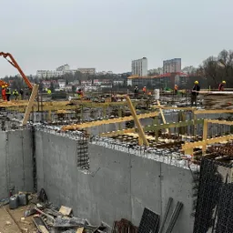 Budowa fundamentów: betonowe ściany szalunkowe z wystającą stalą zbrojeniową, drewniane podpory, pomarańczowa pompa do betonu i pracownicy w żółtych kaskach na tle zabudowy miejskiej w pochmurny...