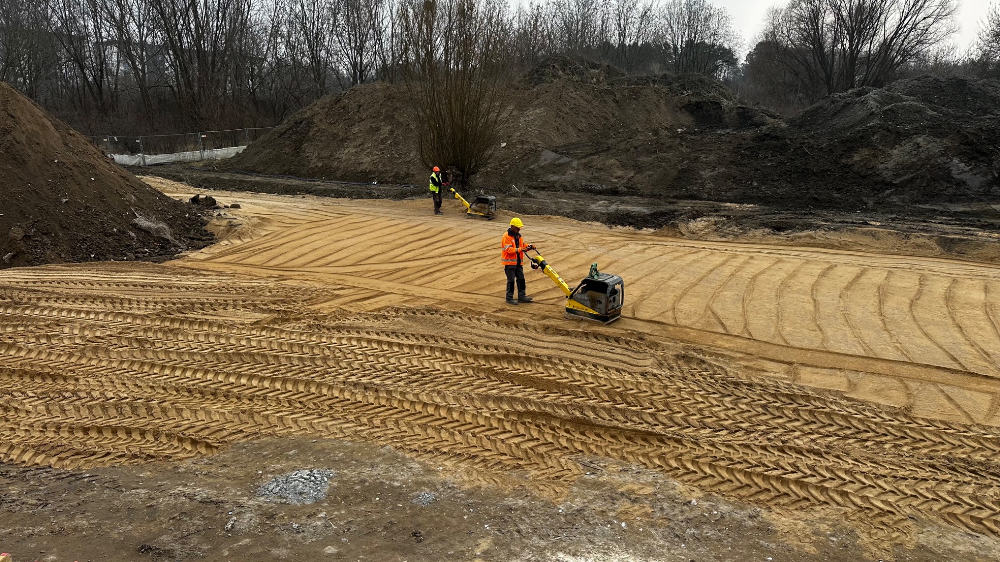 Dwóch robotników w odblaskowych kamizelkach zagęszcza piasek za pomocą zagęszczarek płytowych na placu budowy, w tle hałdy ziemi i nagie drzewa.