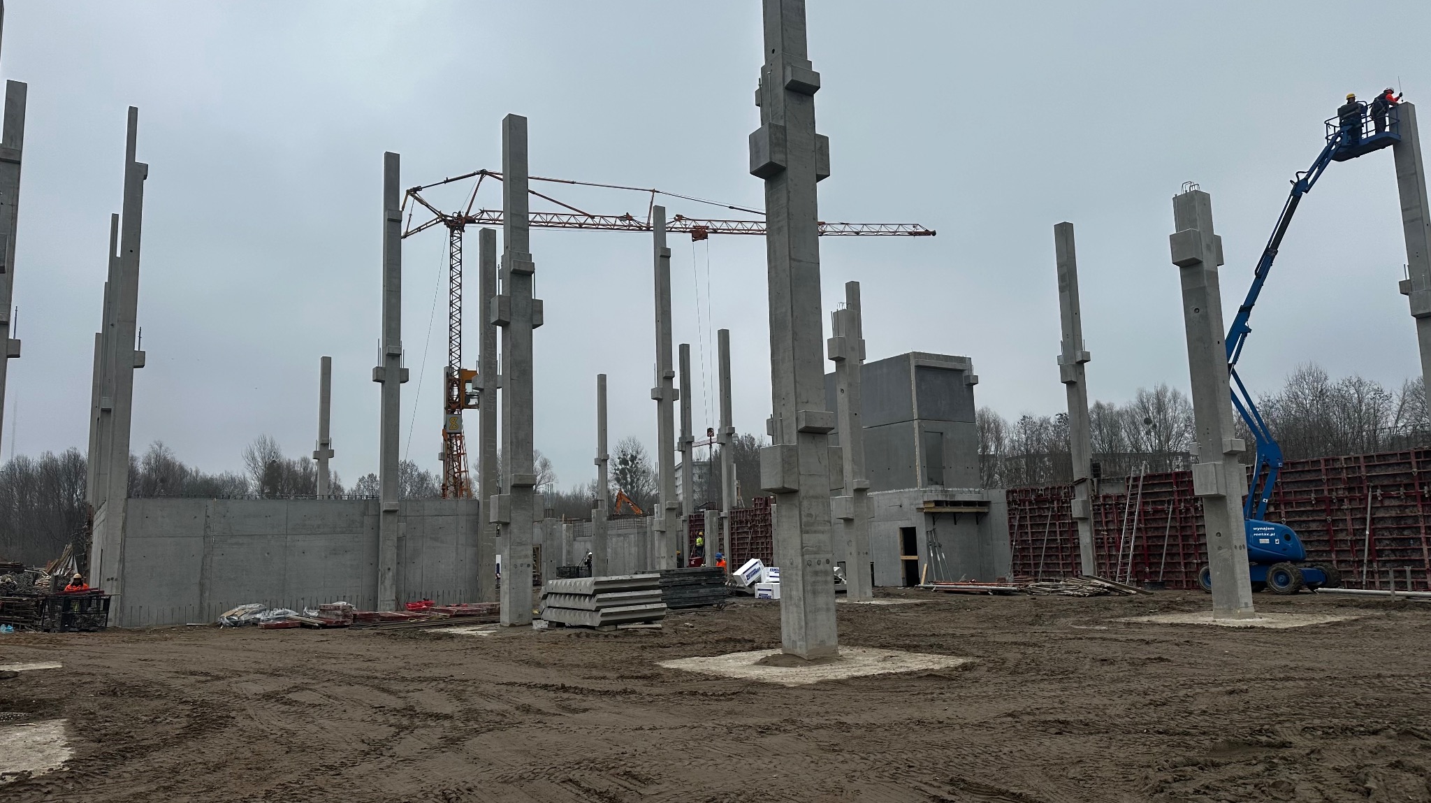 Wznoszenie konstrukcji żelbetowej budynku z użyciem dźwigu i podnośnika koszowego, widoczne betonowe słupy, ściany szalunkowe i zbrojenia.