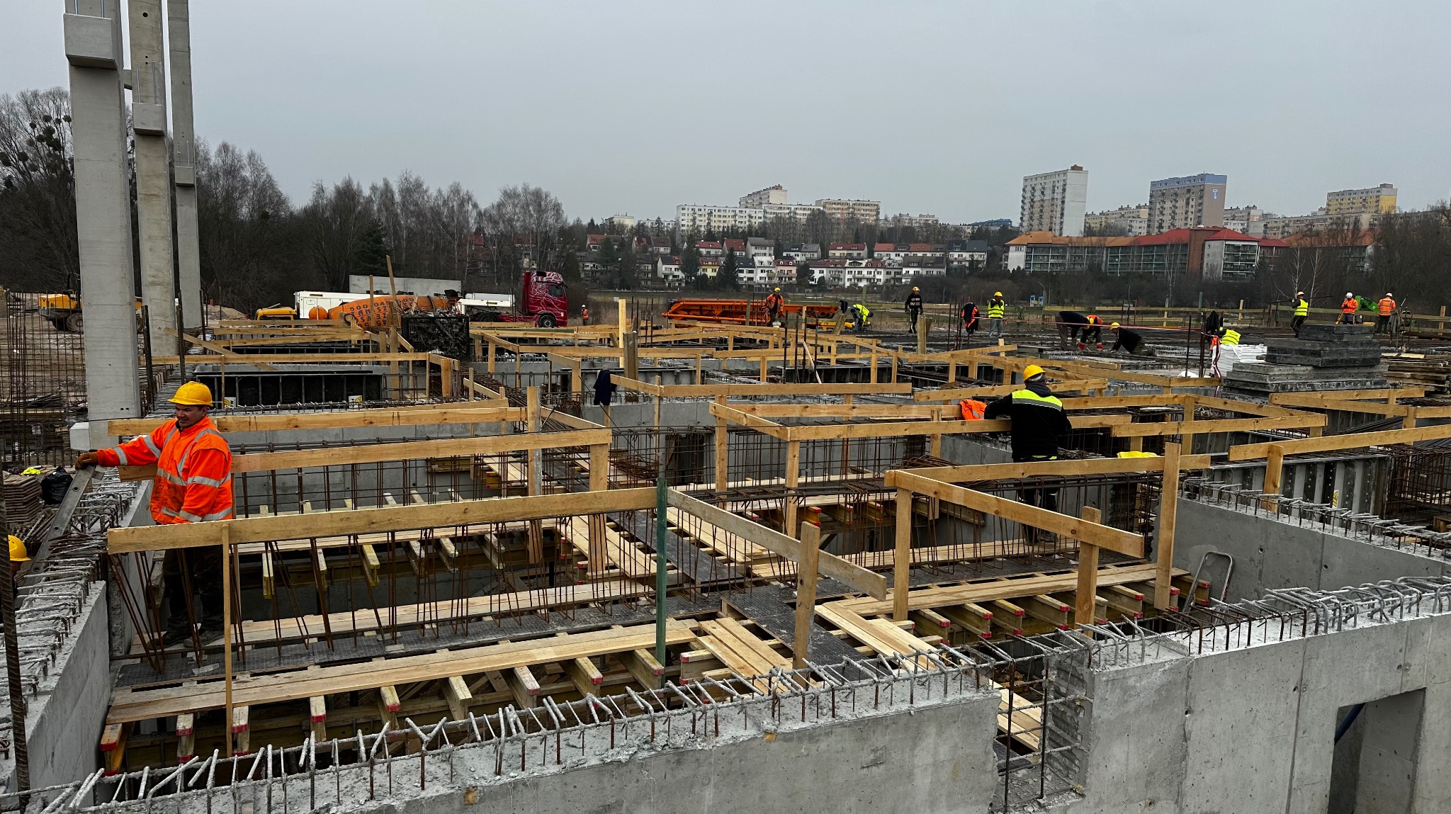Budowa fundamentów z widoczną zbroją i szalunkiem drewnianym, pracownicy w kaskach i kamizelkach odblaskowych w tle, krajobraz miejski w oddali, pochmurne niebo.