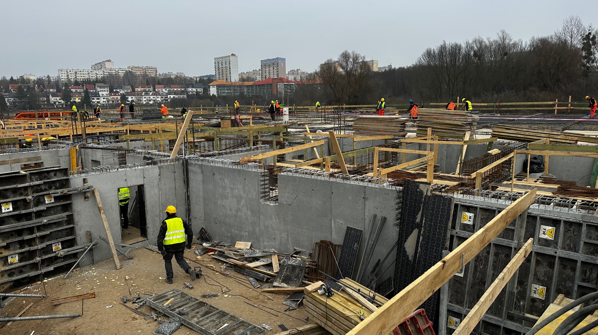 Wykonywanie fundamentów pod budynek wielorodzinny, widoczne szalunki, zbrojenie, betonowe ściany fundamentowe i pracownicy w kaskach i kamizelkach odblaskowych na tle zabudowy miejskiej.