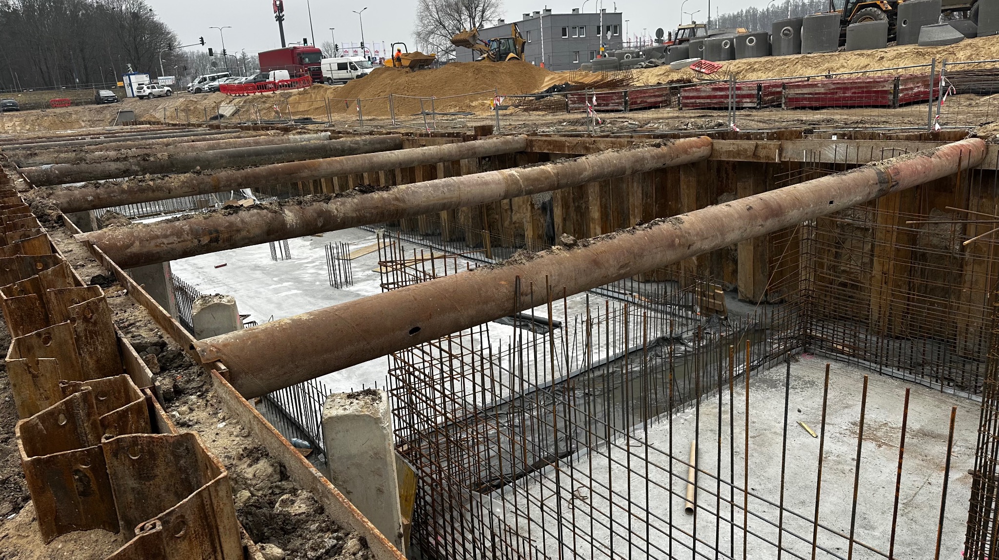 Wykonany wykop zabezpieczony stalowymi rurami i szalunkiem, widoczne zbrojenie fundamentów, w tle koparka na nasypie ziemi i budynki.