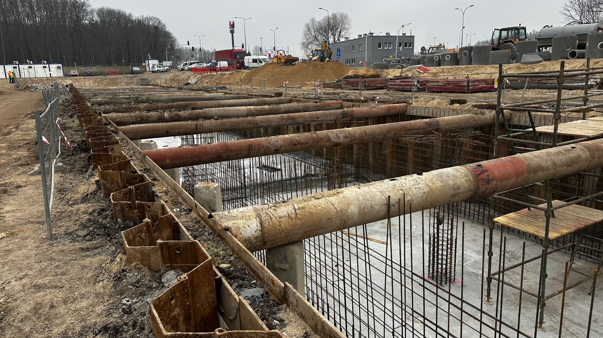 Szeroki wykop zabezpieczony metalowymi szalunkami i rurami, z widoczną siatką zbrojeniową, na tle placu budowy z maszynami i elementami betonowymi.