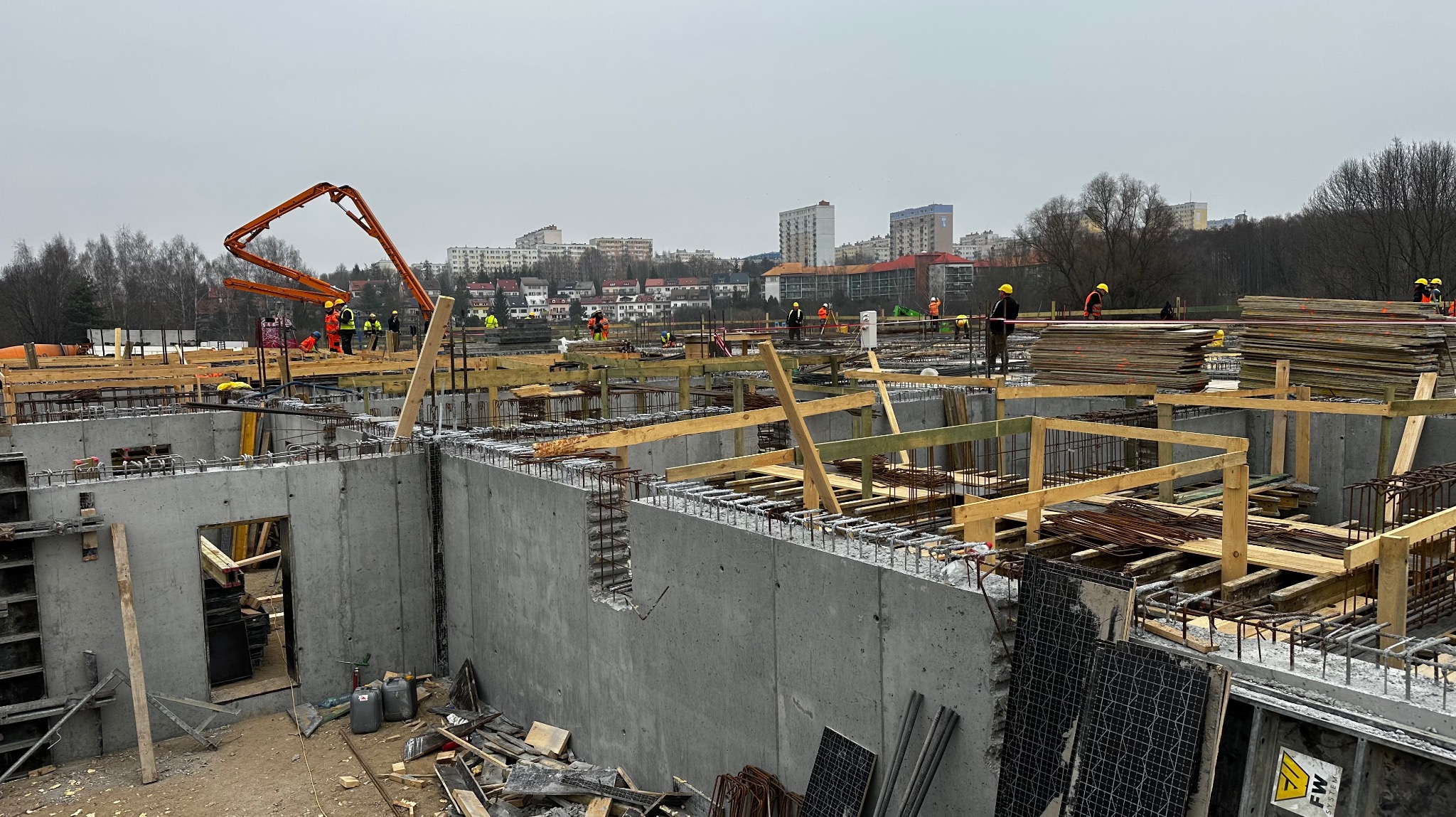Budowa fundamentów: betonowe ściany szalunkowe z wystającą stalą zbrojeniową, drewniane podpory, pomarańczowa pompa do betonu i pracownicy w żółtych kaskach na tle zabudowy miejskiej w pochmurny...