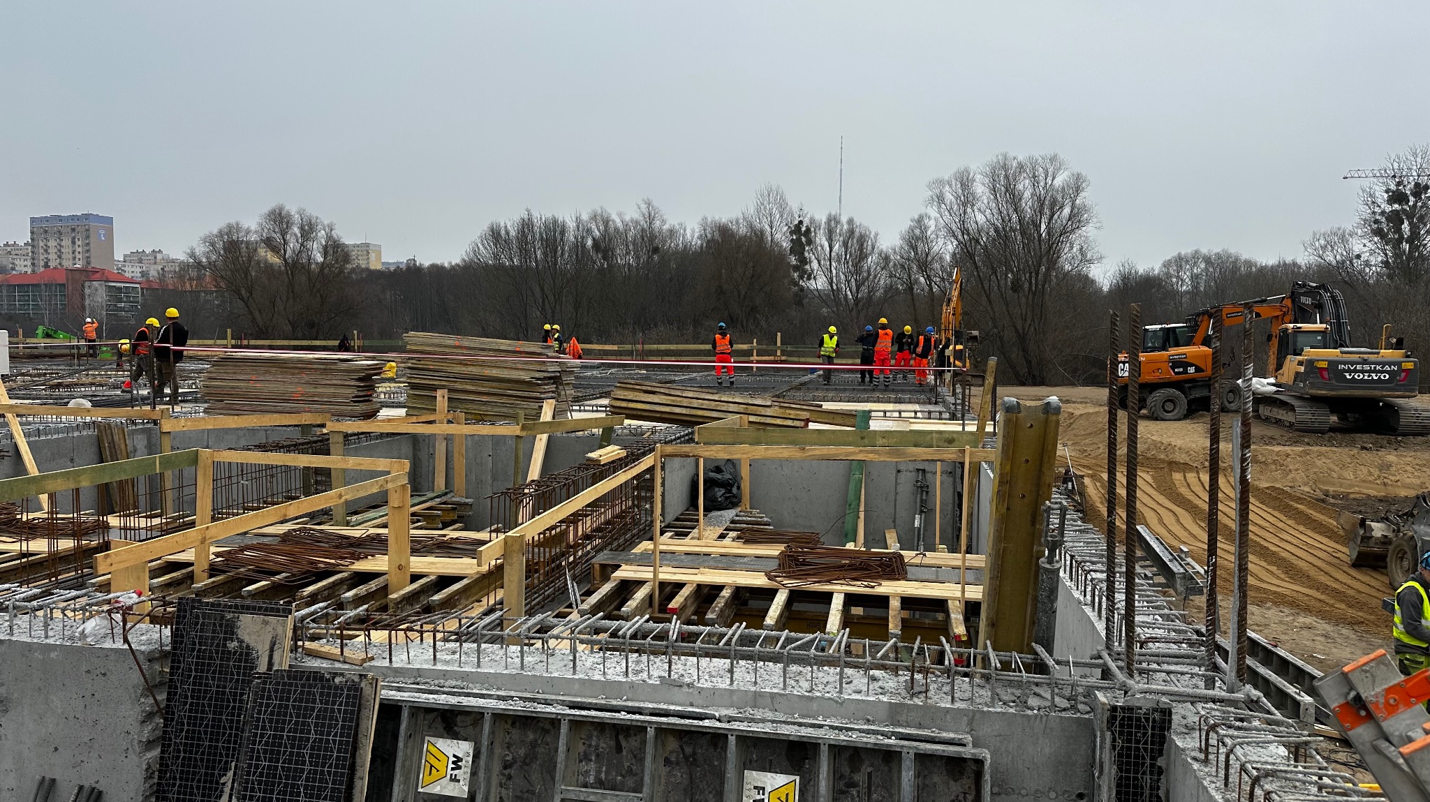 Budowa fundamentów: widok na szalunki, zbrojenia i ekipę budowlaną w kaskach, z dwoma koparkami w tle, na tle drzew i budynków mieszkalnych w pochmurny dzień.